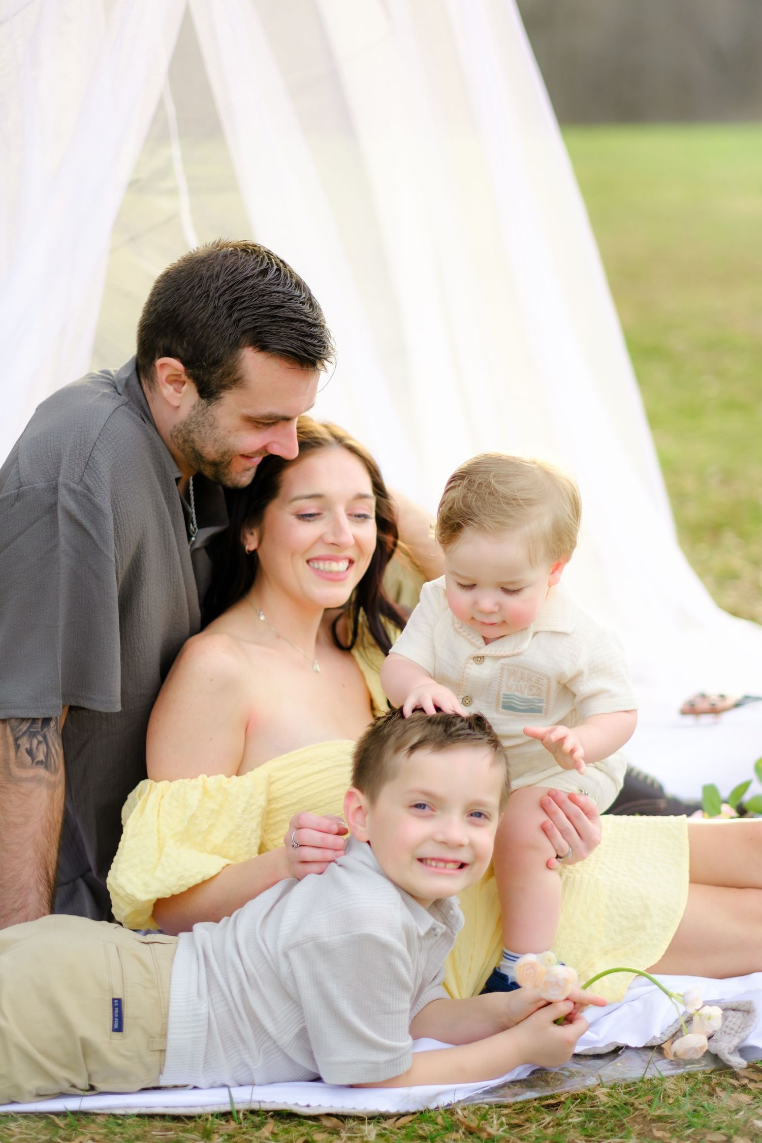 Happy family of four sitting outdoors on a blanket near a white canopy tent, enjoying a warm sunny day together.