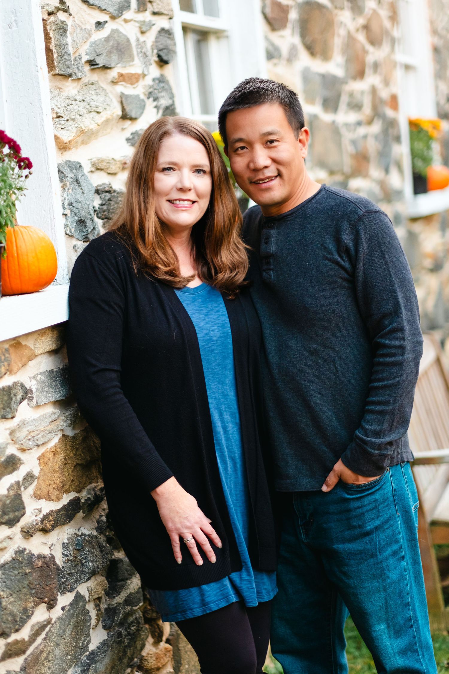 A smiling couple poses against a rustic stone wall with fall pumpkins and flowers in the background.
