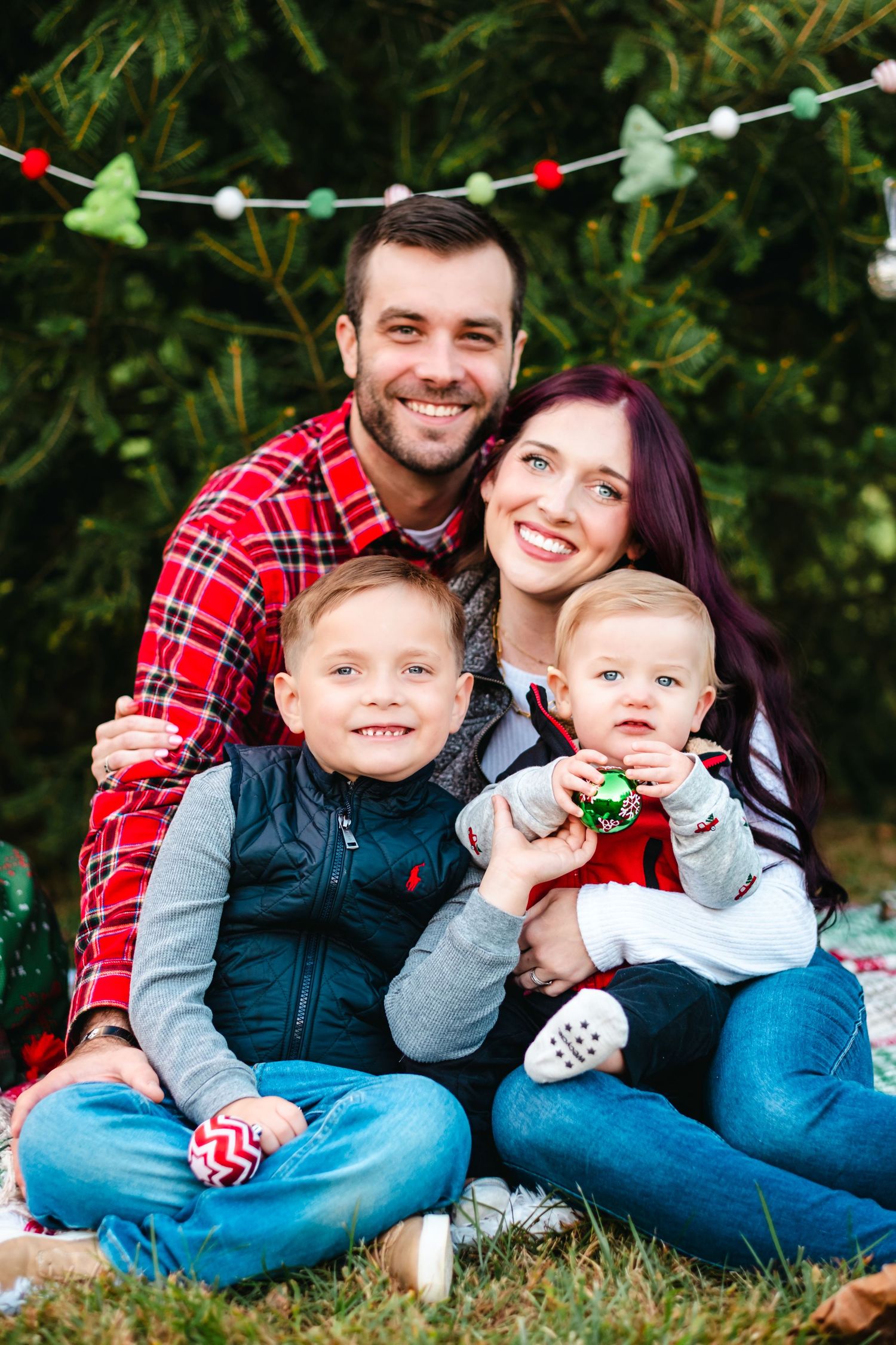 Family of four posing outdoors near Christmas trees with holiday decorations, parents smiling with two young boys.
