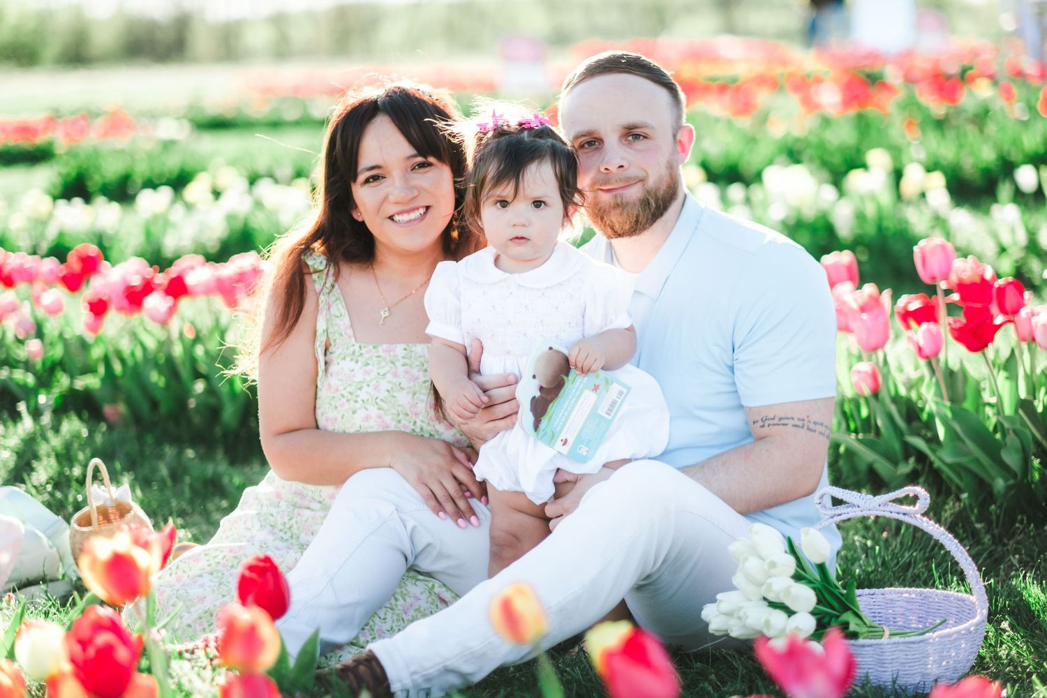 Family of three sitting among red and pink tulips in a flower field, smiling couple holding baby girl in white outfit.