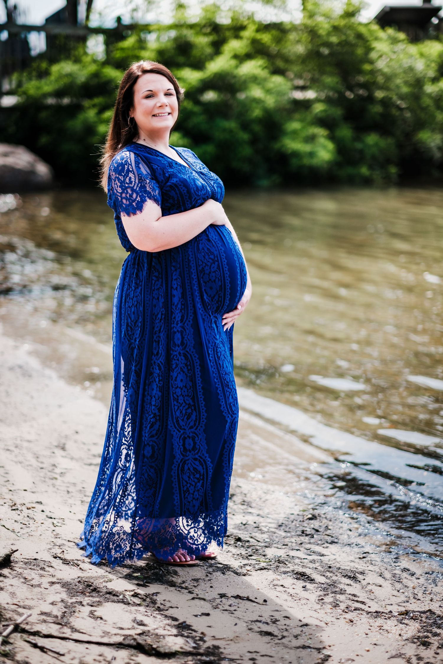 Smiling pregnant woman in blue lace maxi dress standing by a river shore, cradling her baby bump outdoors.