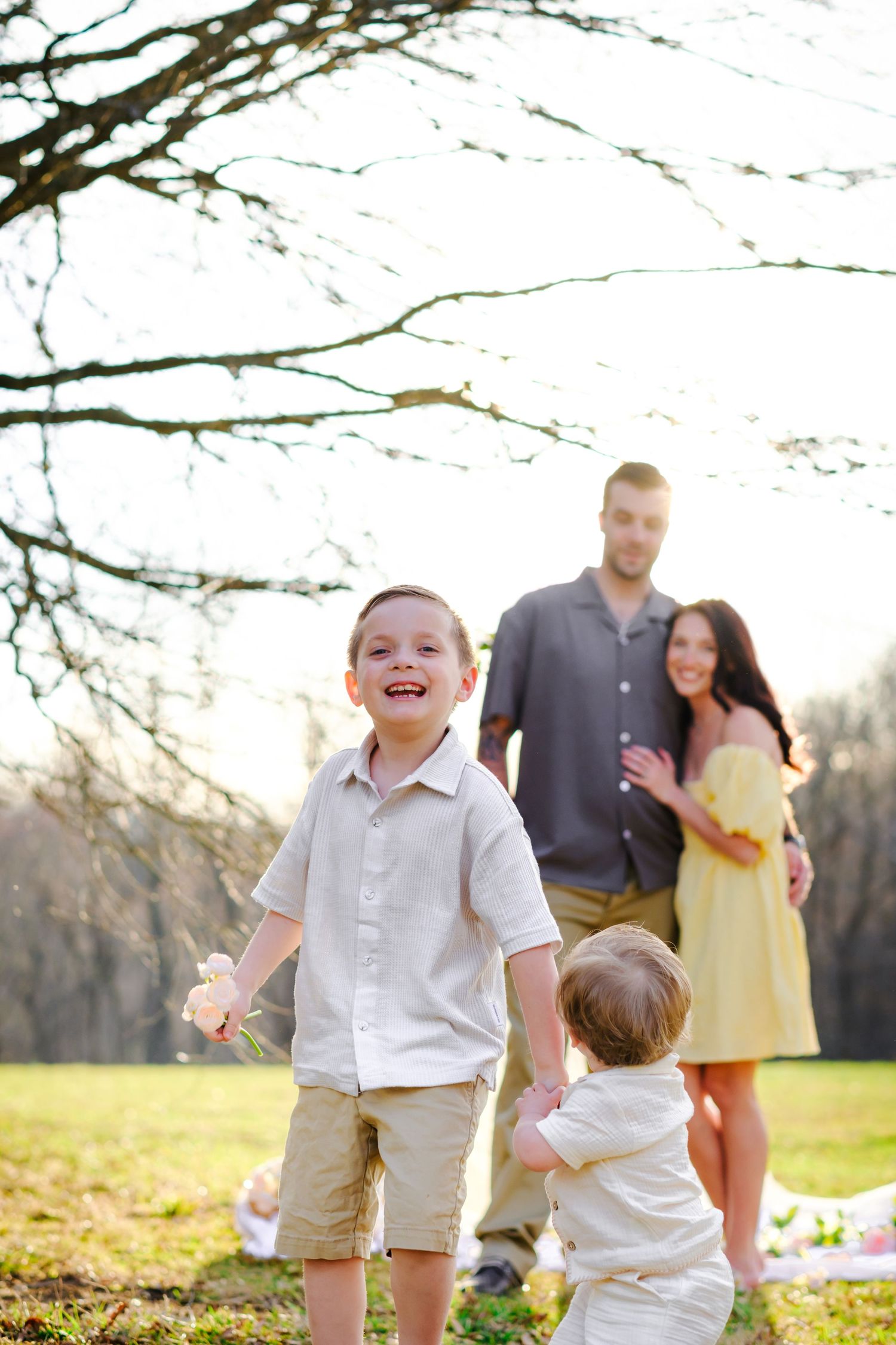 Young boy laughs outdoors as parents stand under tree watching their two sons play during family session.