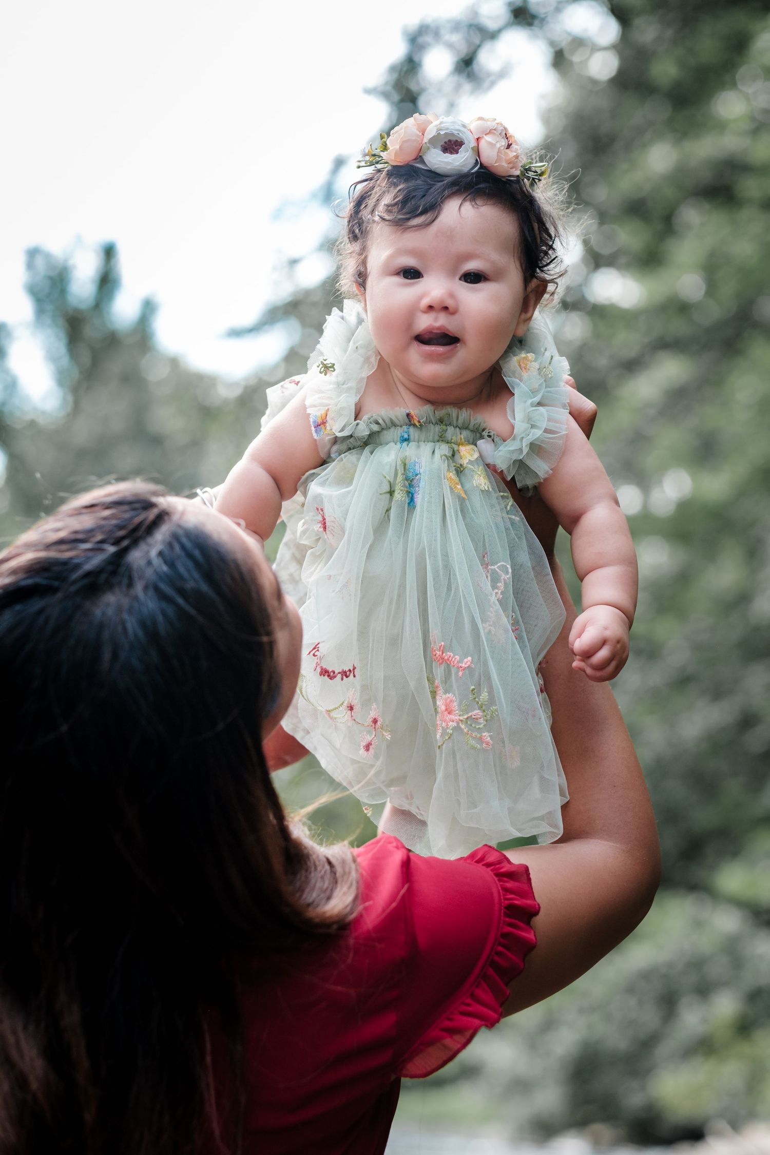 A mother in a red dress lifts her baby girl wearing a floral tulle dress and flower crown outdoors.