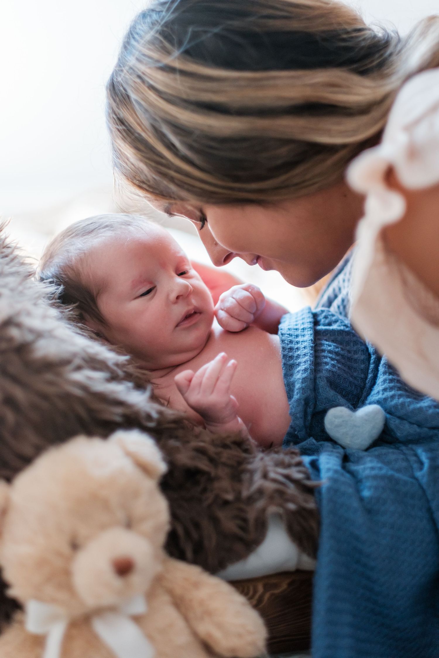 Mother tenderly nose-touching her newborn baby wrapped in blue blanket beside a teddy bear.
