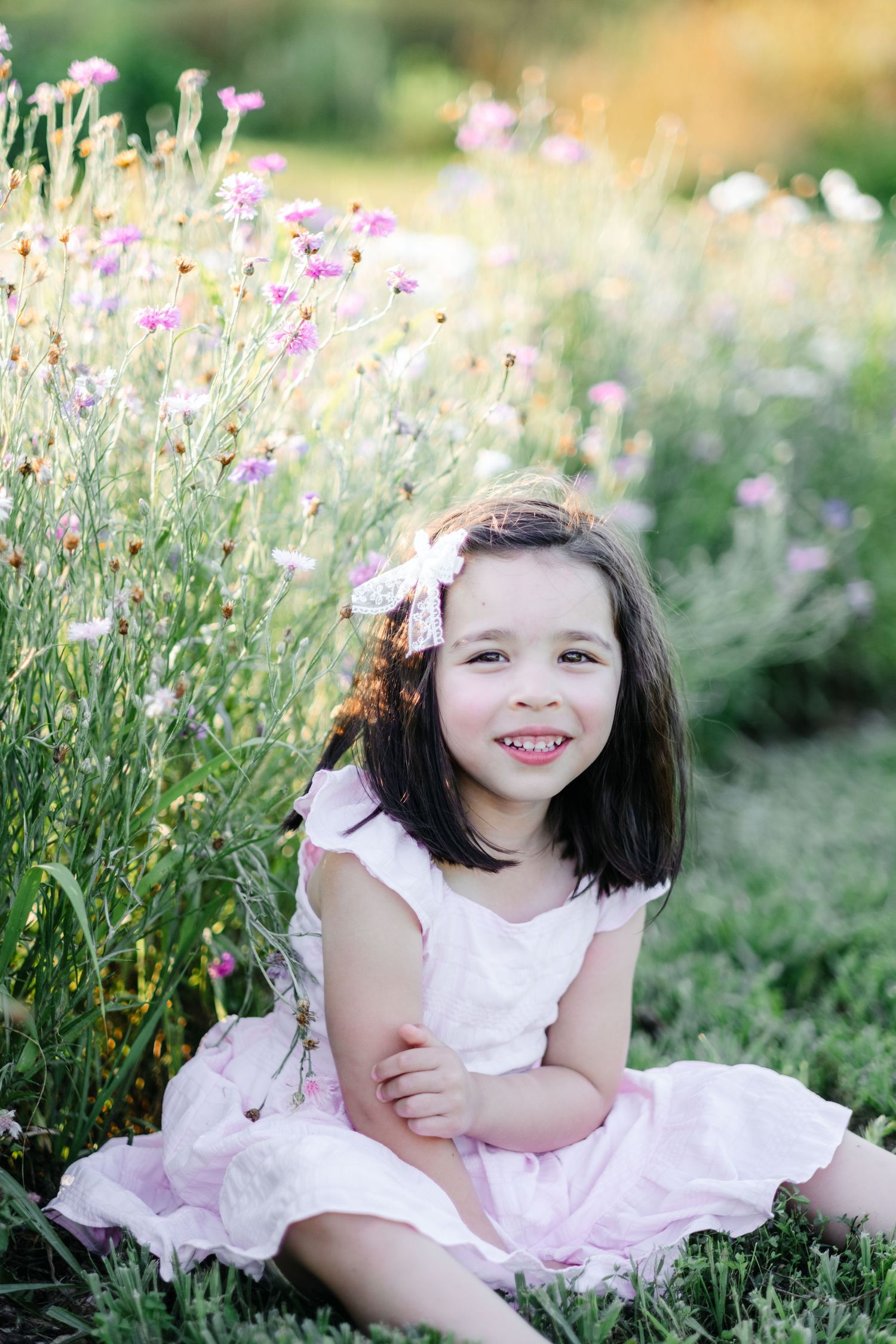 Young girl in pink dress sitting among wildflowers, smiling with a bow in her dark hair on a sunny day.
