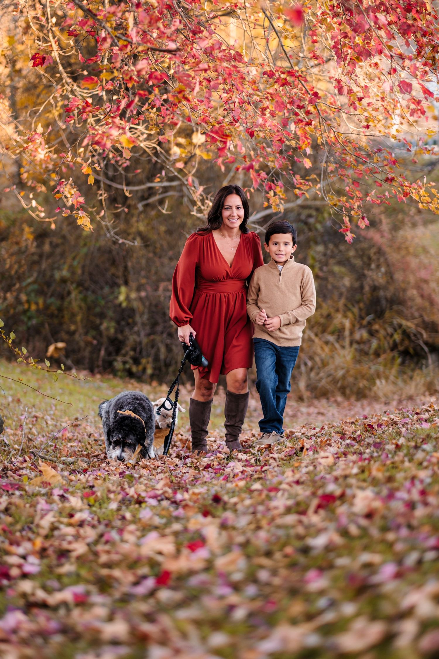 Woman in red dress and boy walk dog through colorful autumn forest path covered in fallen leaves.