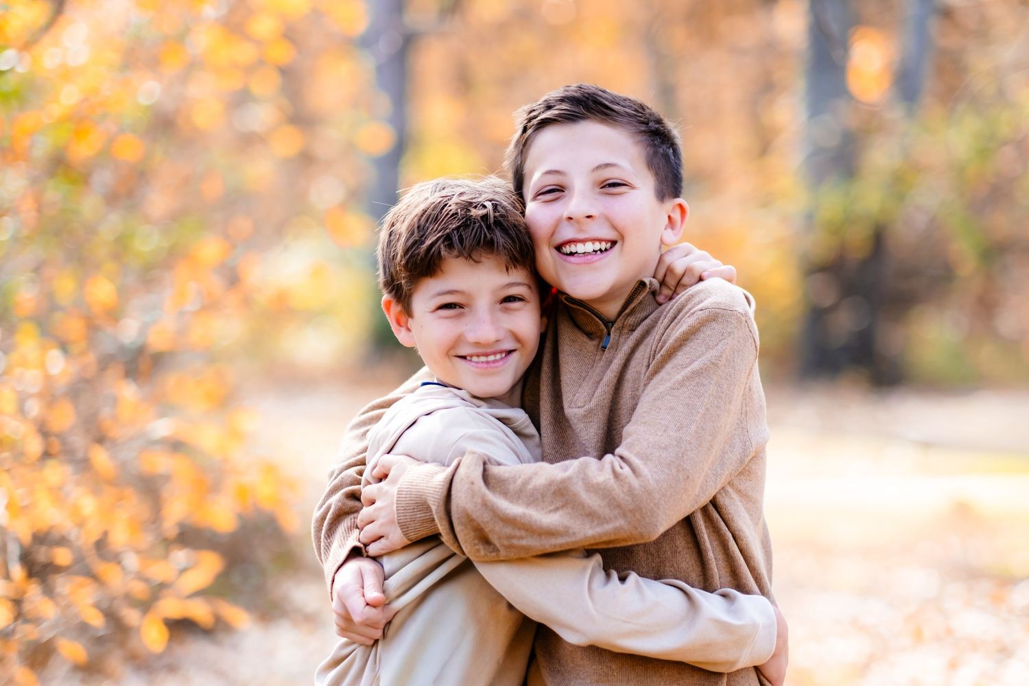 Two smiling boys hugging outdoors in autumn forest with golden foliage, wearing beige hoodies on a nature path.