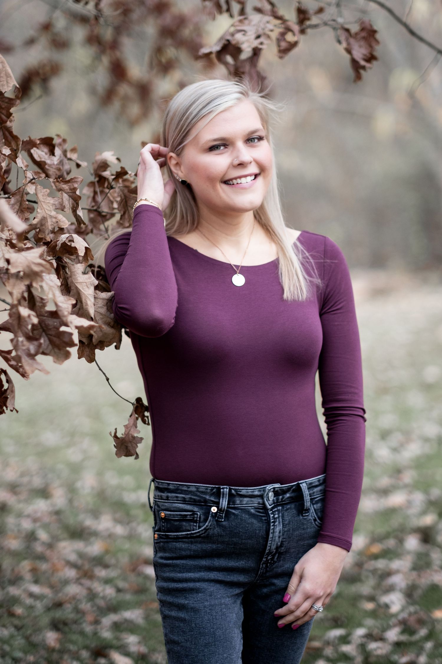 Smiling blonde woman in burgundy long-sleeve top and jeans poses outdoors among autumn oak leaves.
