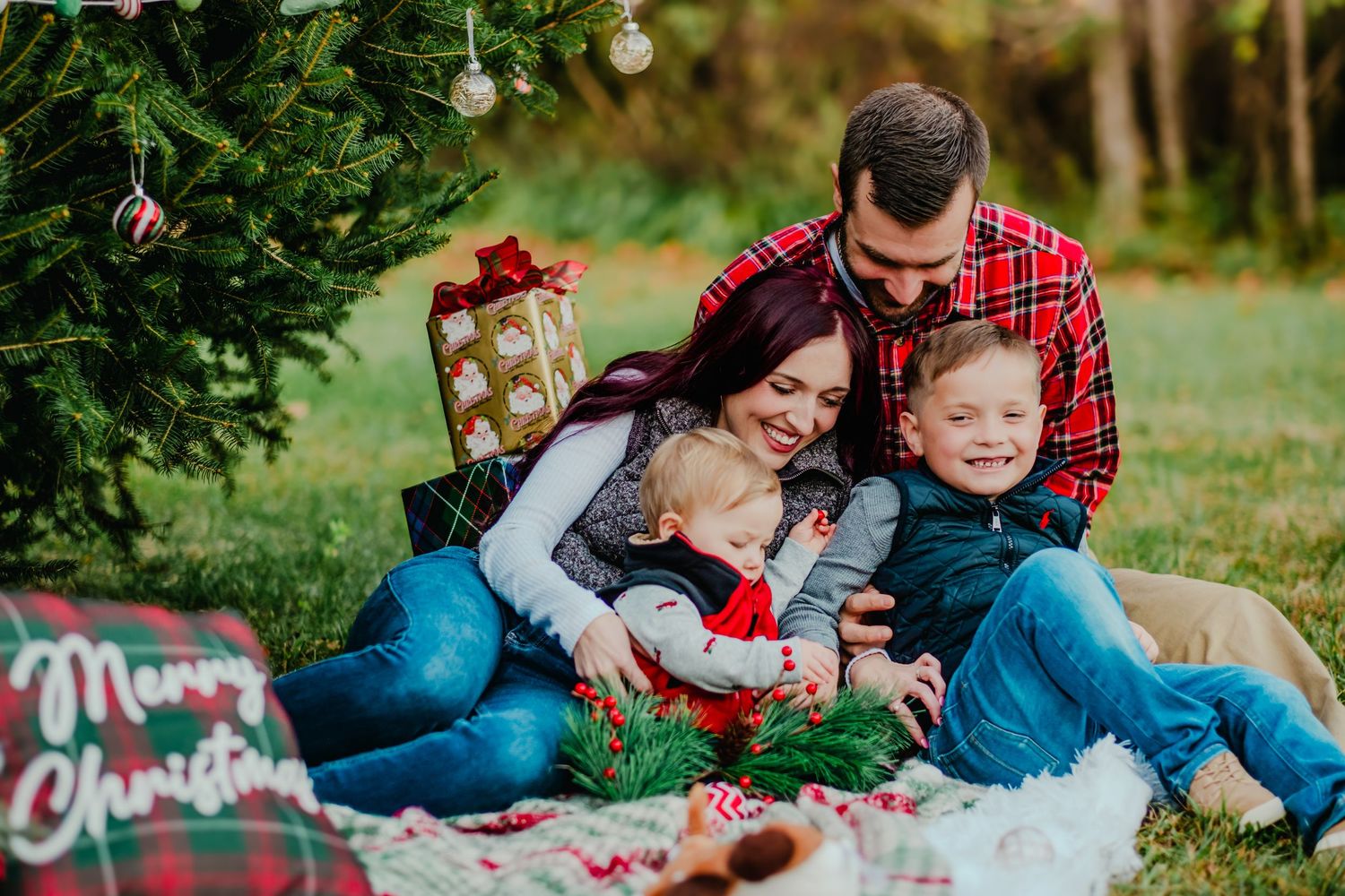 Family of four sitting on a Christmas blanket outdoors, smiling and embracing near a decorated holiday tree.