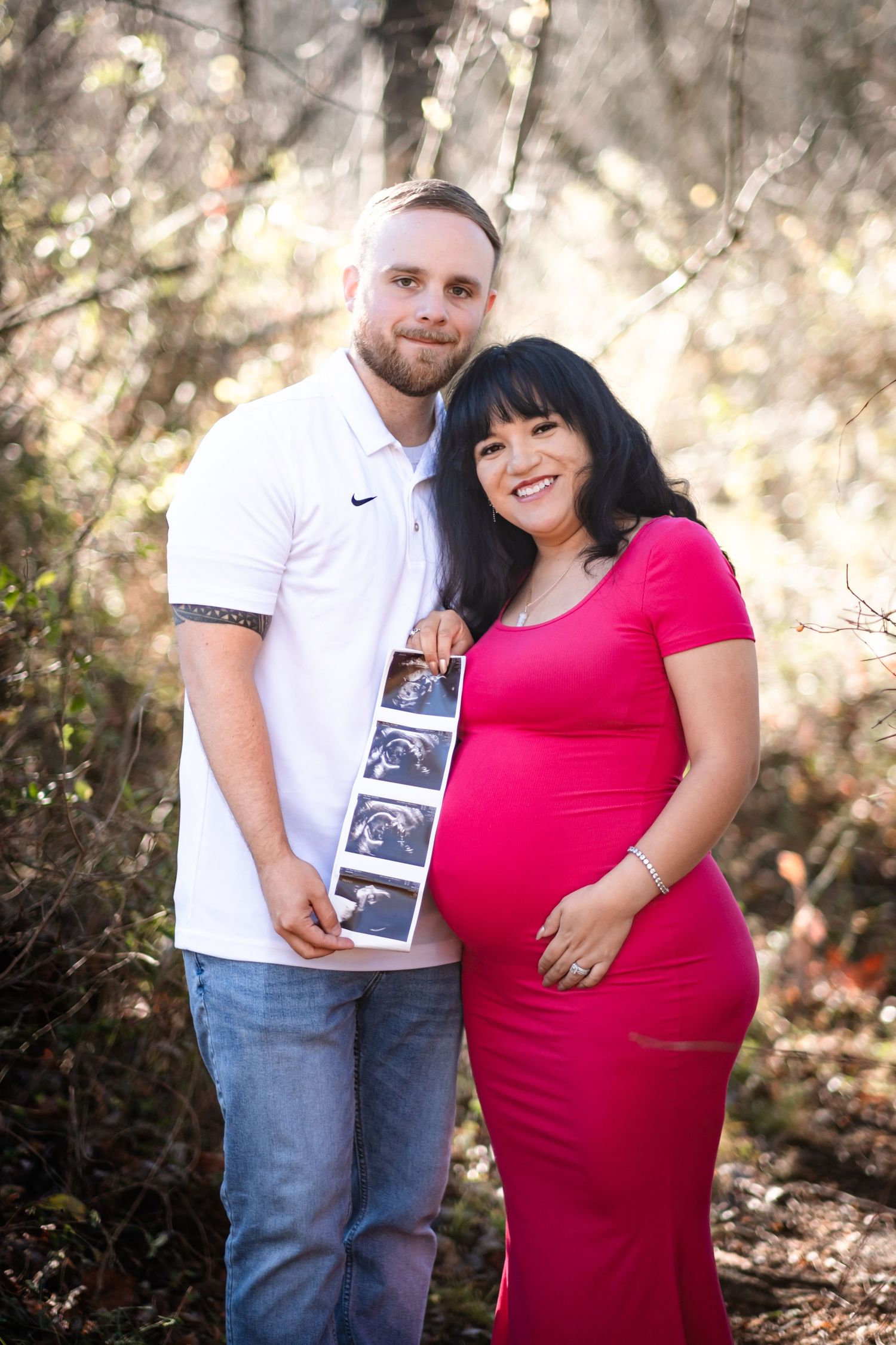 Smiling pregnant couple holding ultrasound photos outdoors, woman in red dress cradling baby bump, man in white polo shirt.