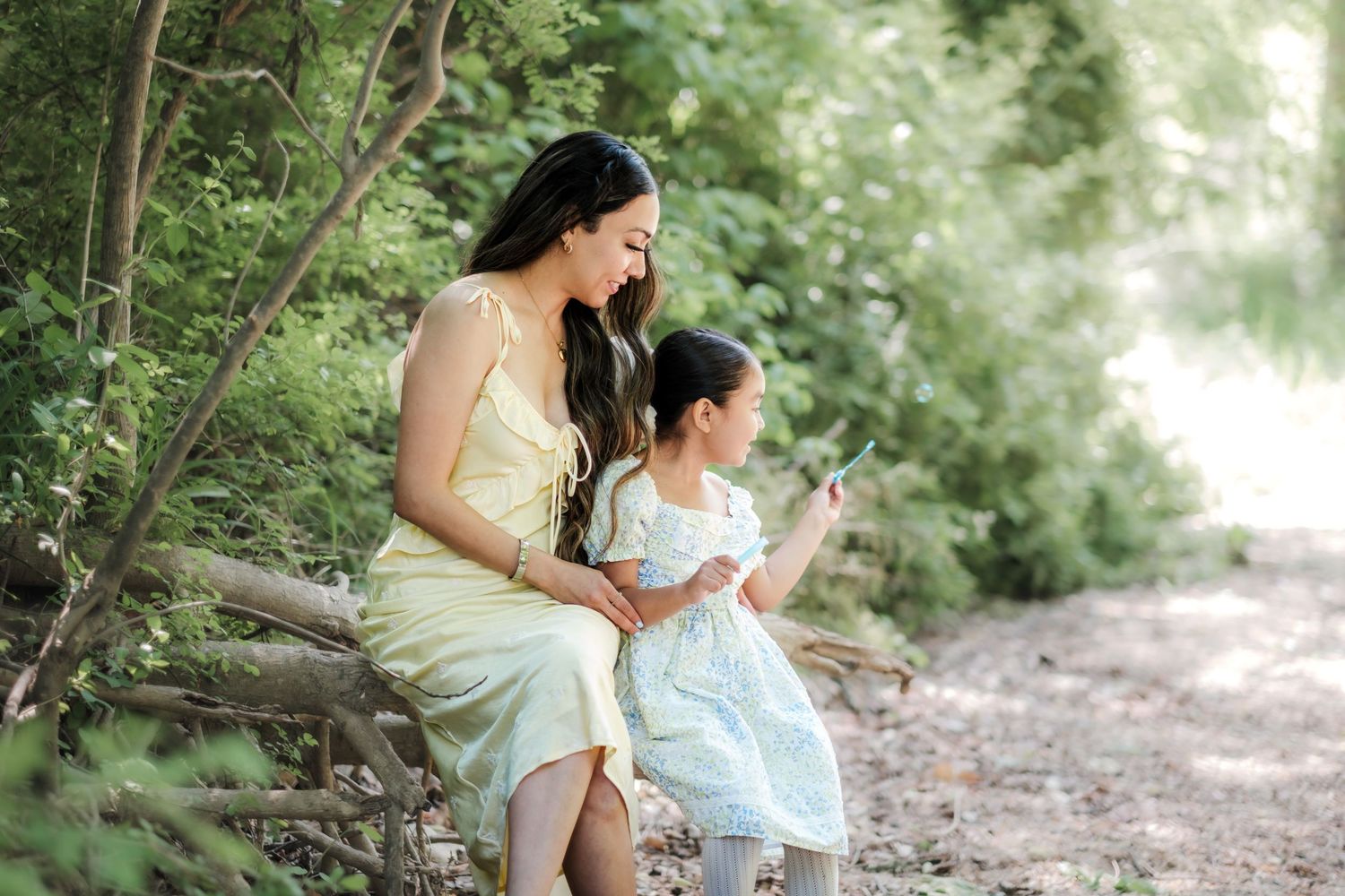 Two figures in light dresses sit together on a forest path surrounded by lush greenery on a sunny day.