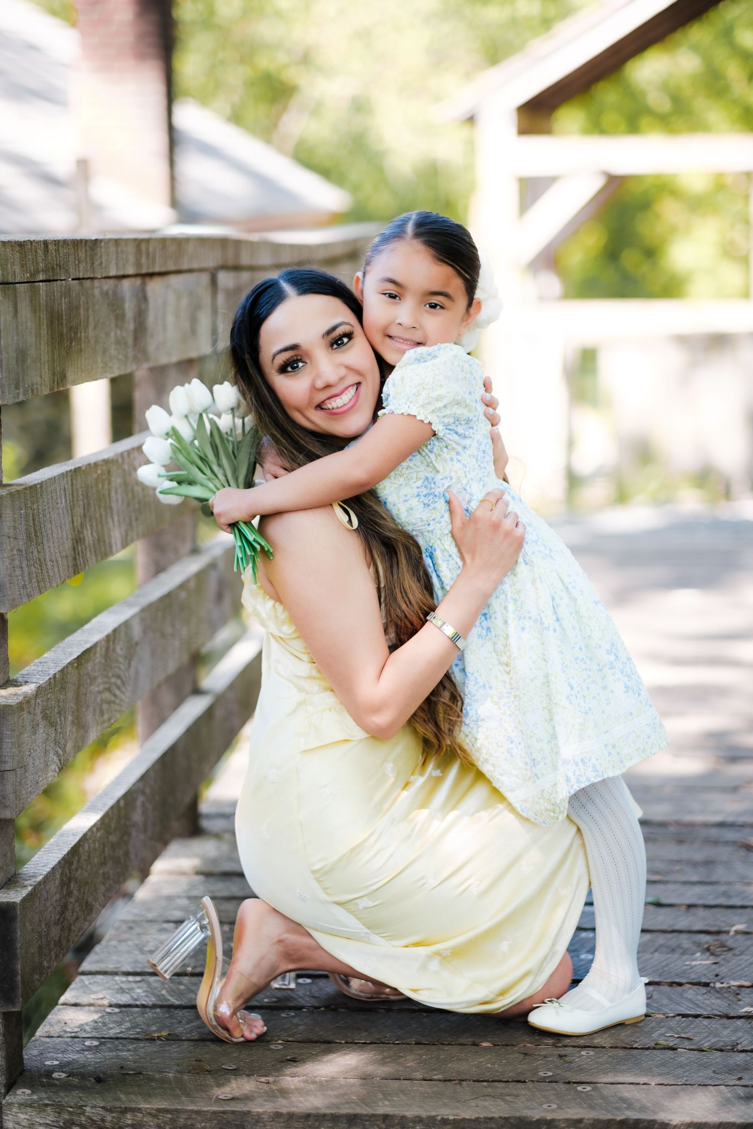A smiling mother in a yellow dress hugs her daughter on a wooden bridge while holding white tulips.