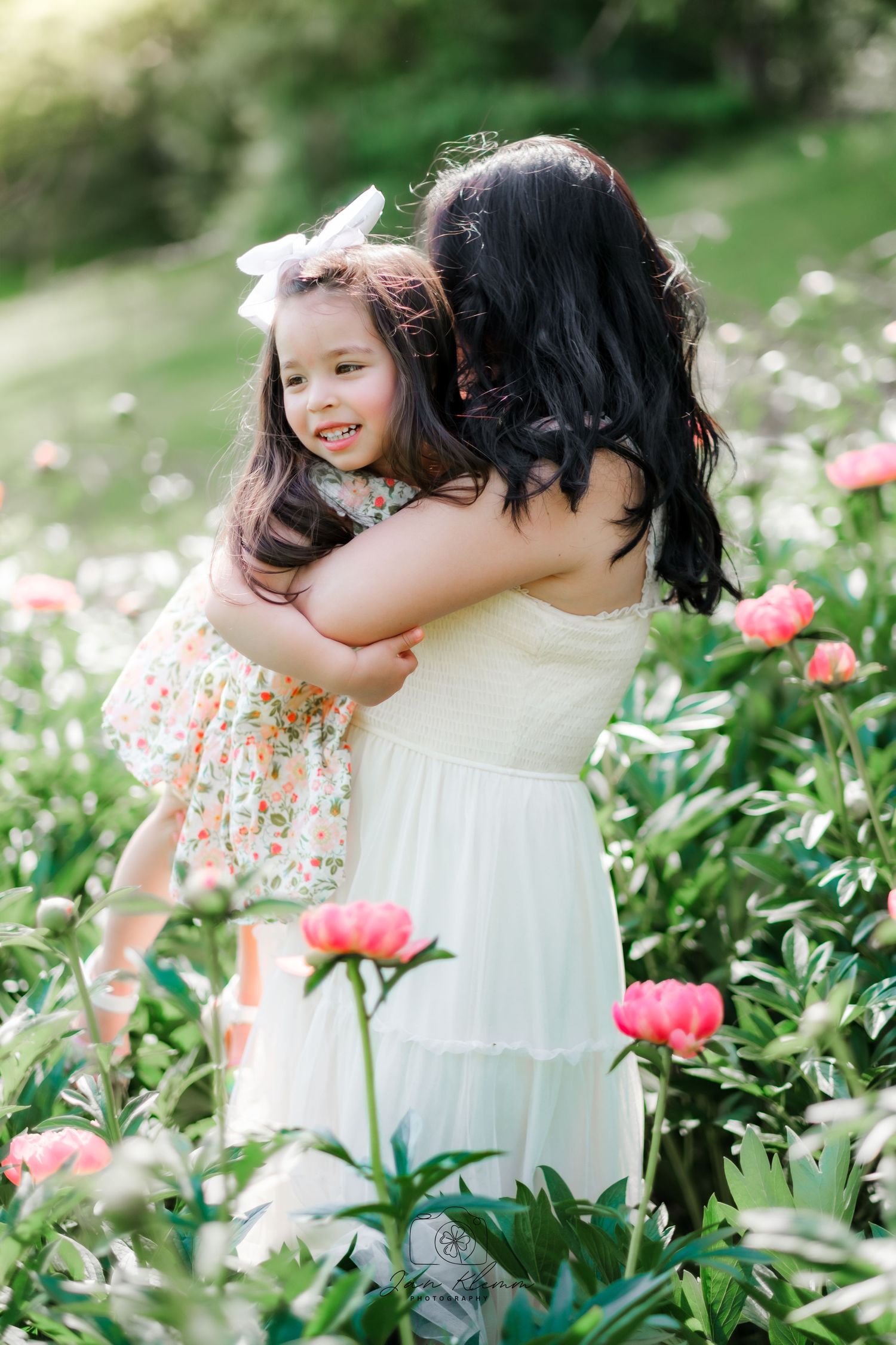Mother in white dress hugging smiling little girl with bow in blooming peony garden.