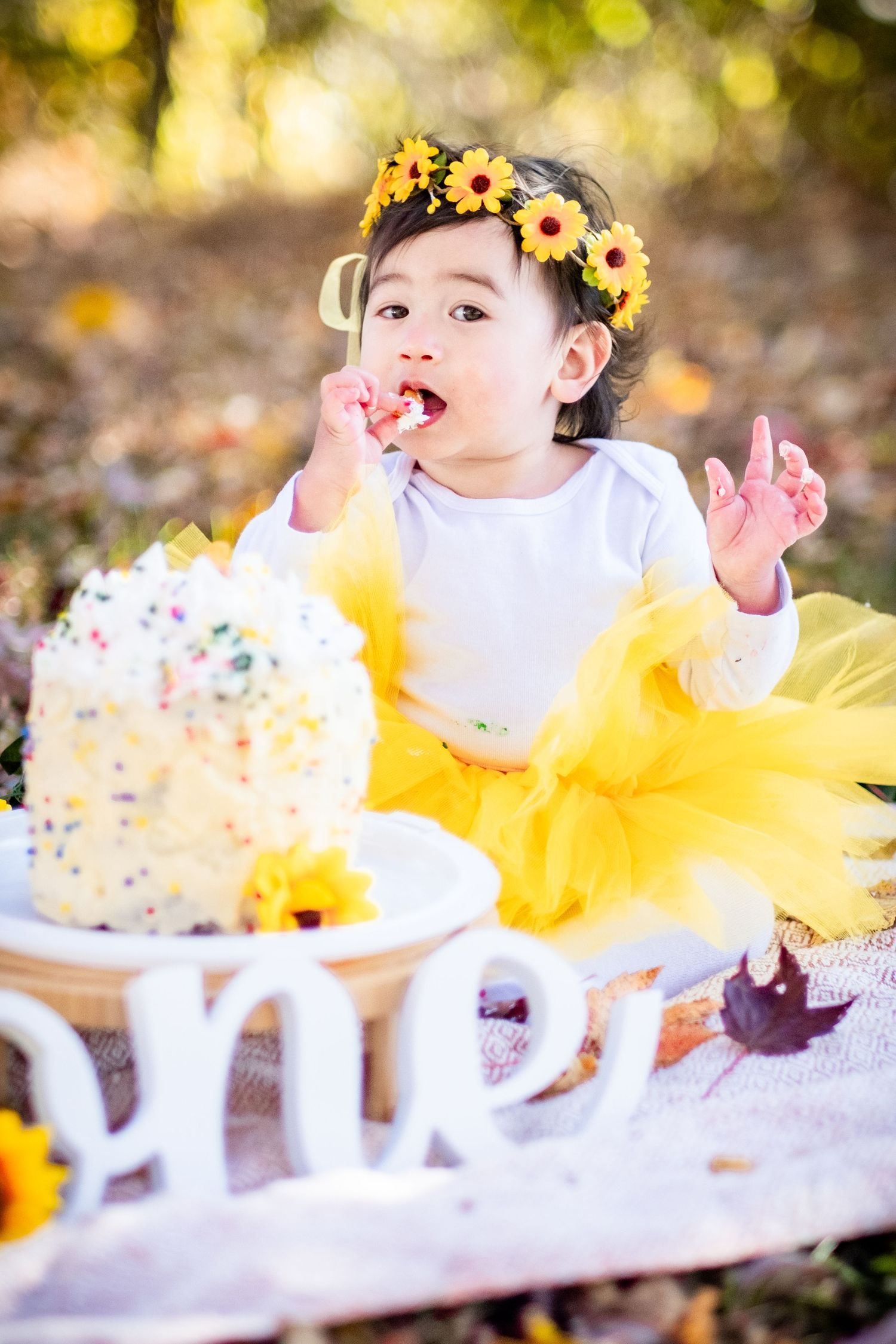 Baby girl in yellow tutu and sunflower crown eating first birthday smash cake outdoors.