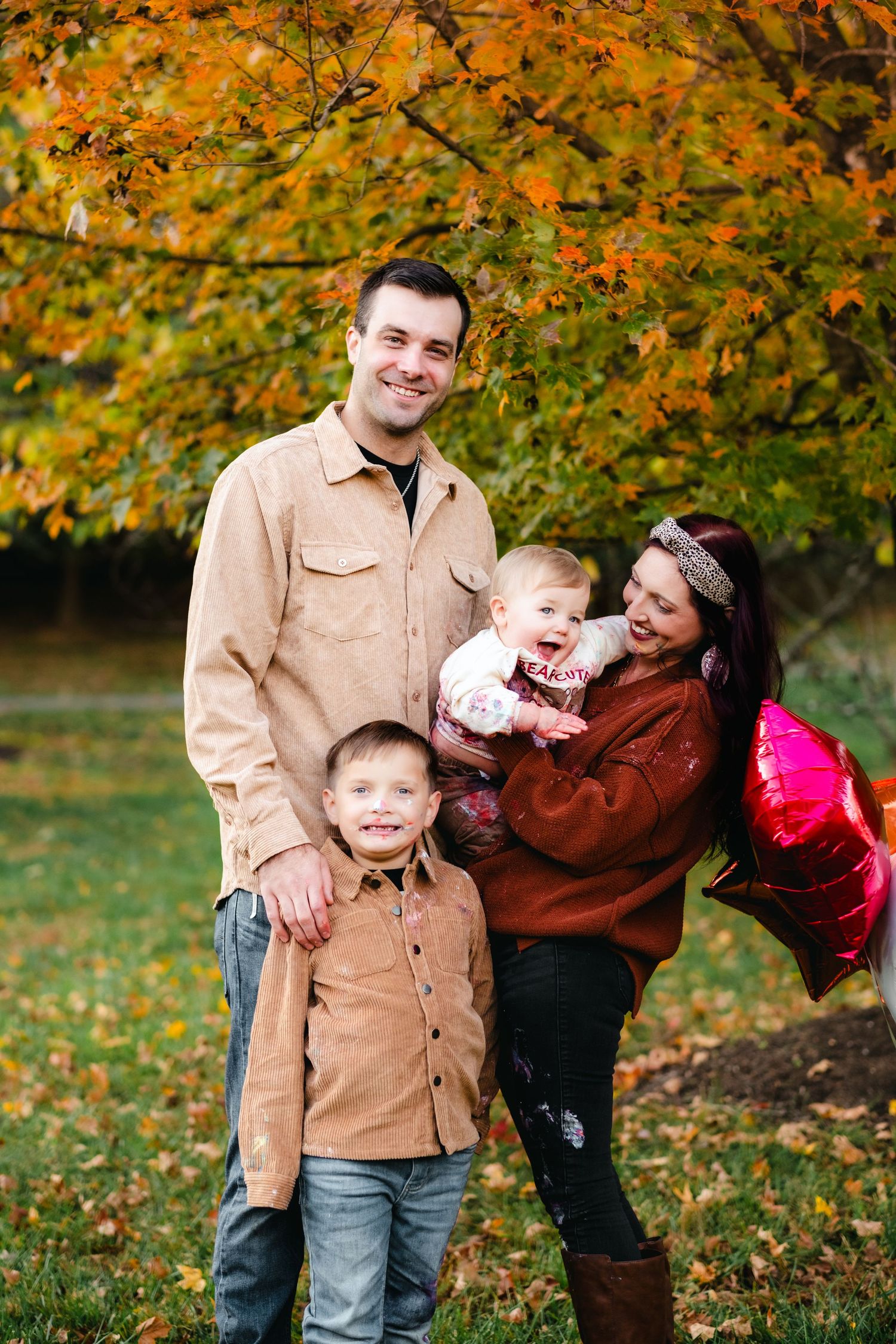 Happy family of four posing outdoors in autumn with vibrant orange foliage and pink balloons.