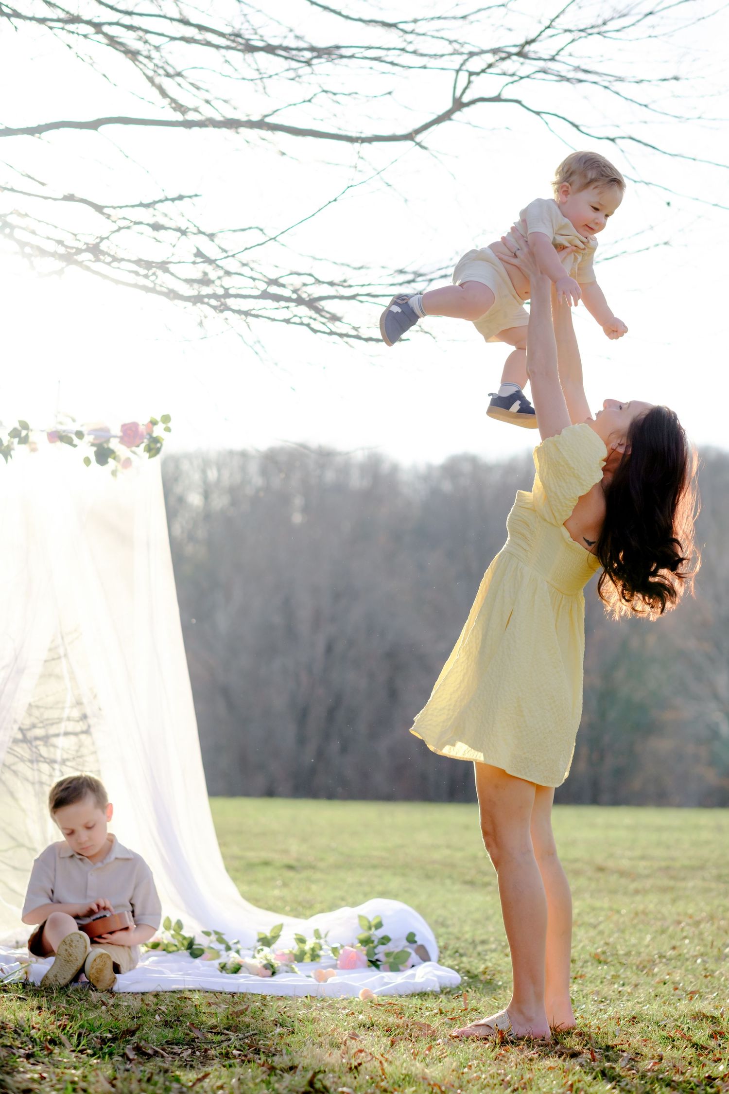 A joyful mother in a yellow dress lifts her baby high in the air outdoors while her young son plays nearby.