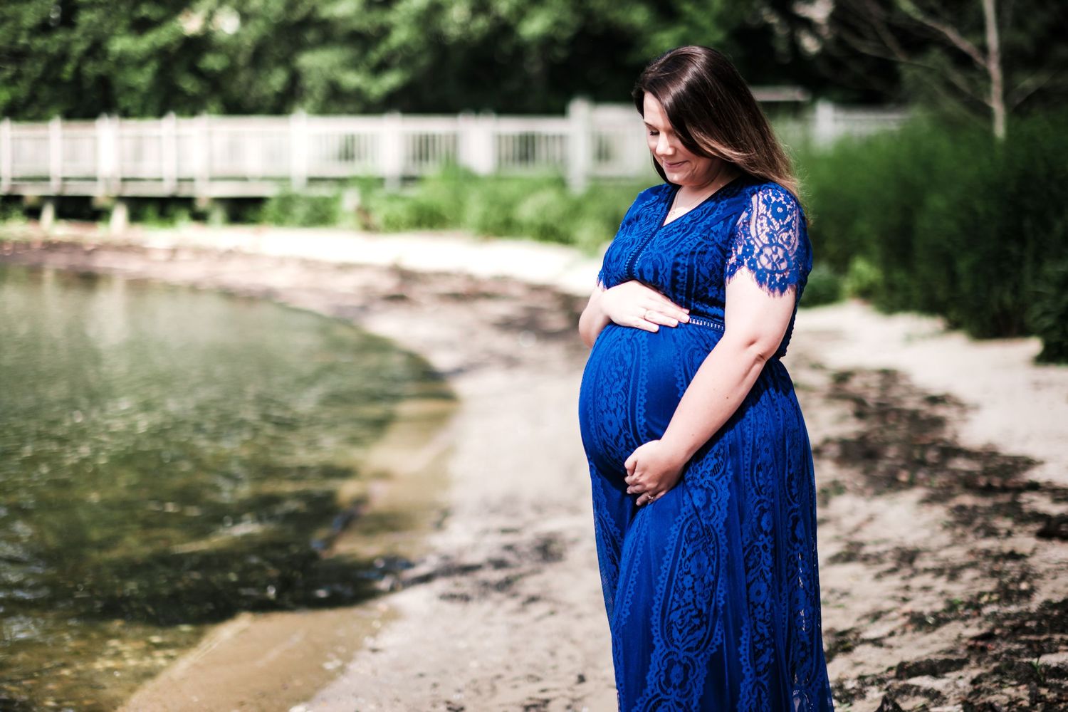 Pregnant woman in blue lace dress holding belly on sandy shore near calm water.