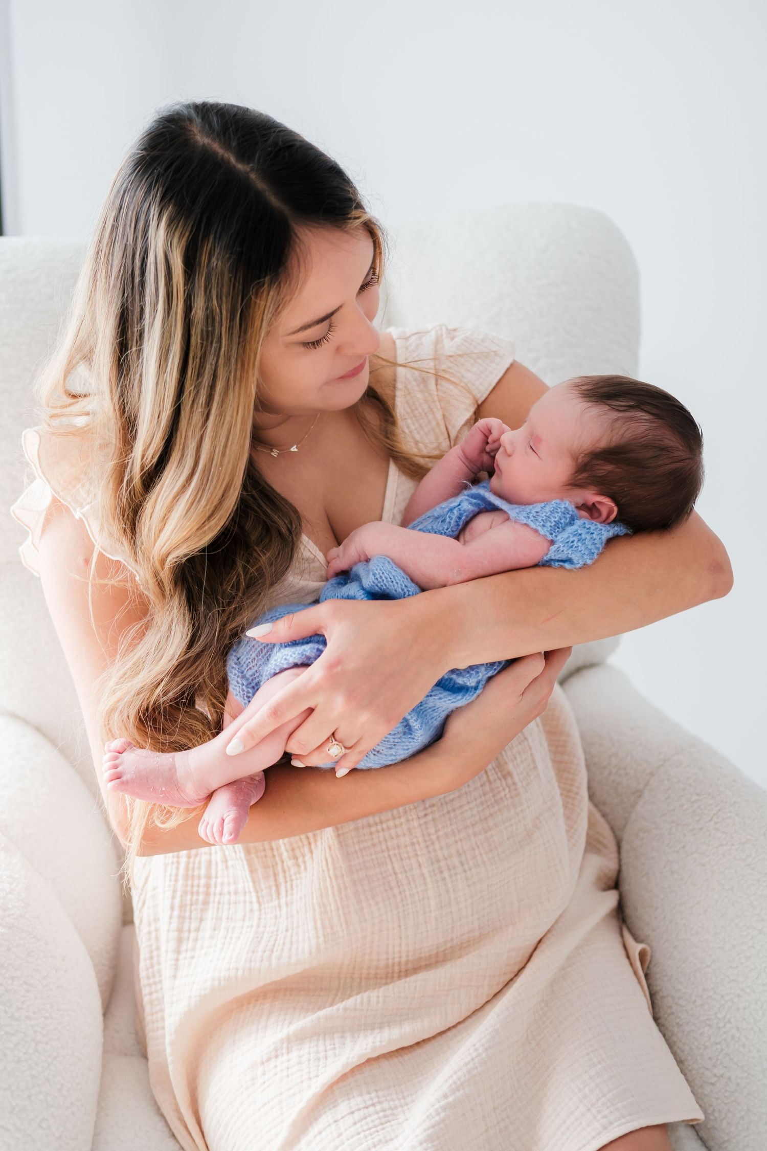 A young mother in a cream dress tenderly holds her sleeping newborn wrapped in a blue knit blanket in a white chair.