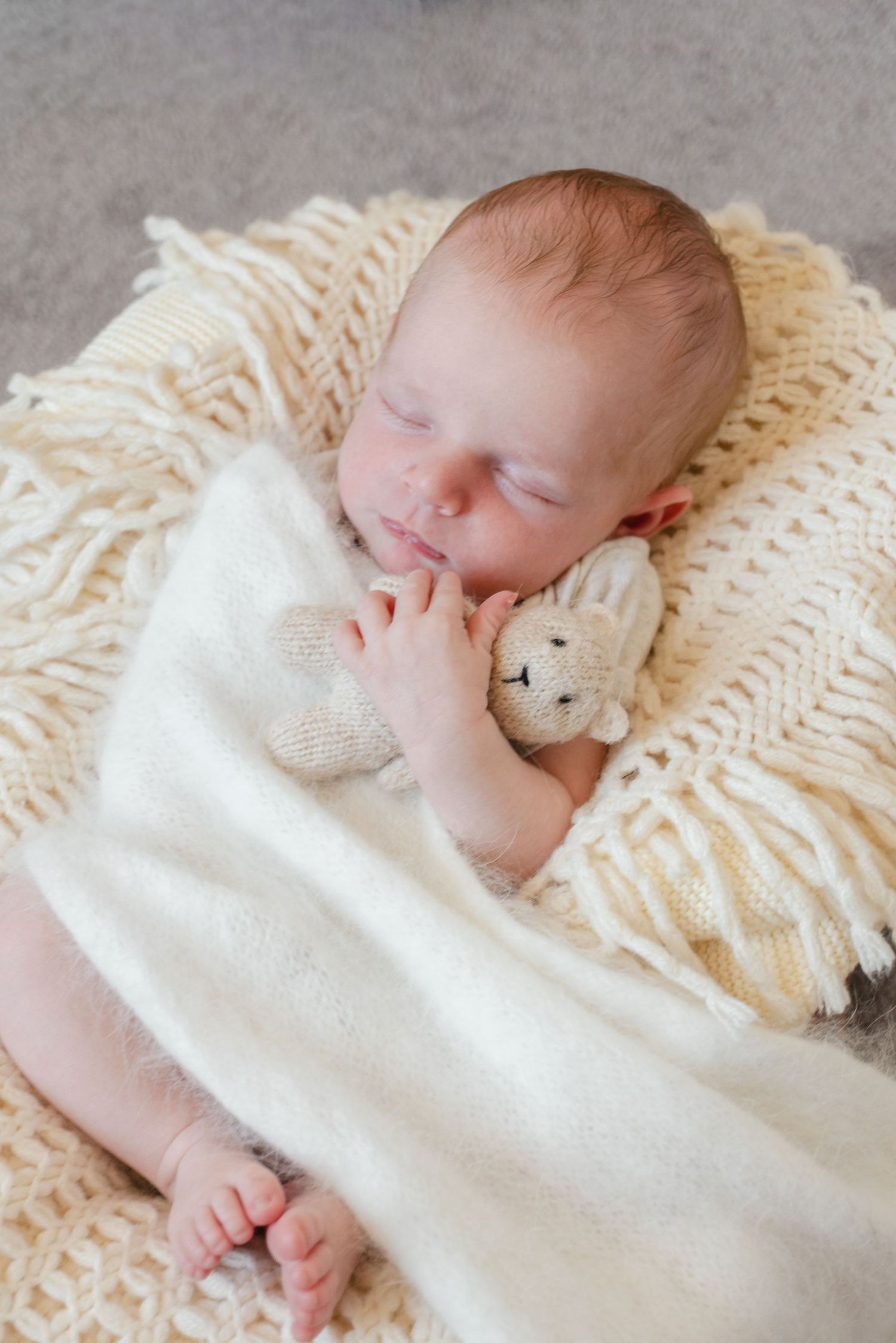 Peaceful newborn baby sleeping on cream knit blanket, wrapped in soft white wrap, holding a tiny knitted bunny toy.