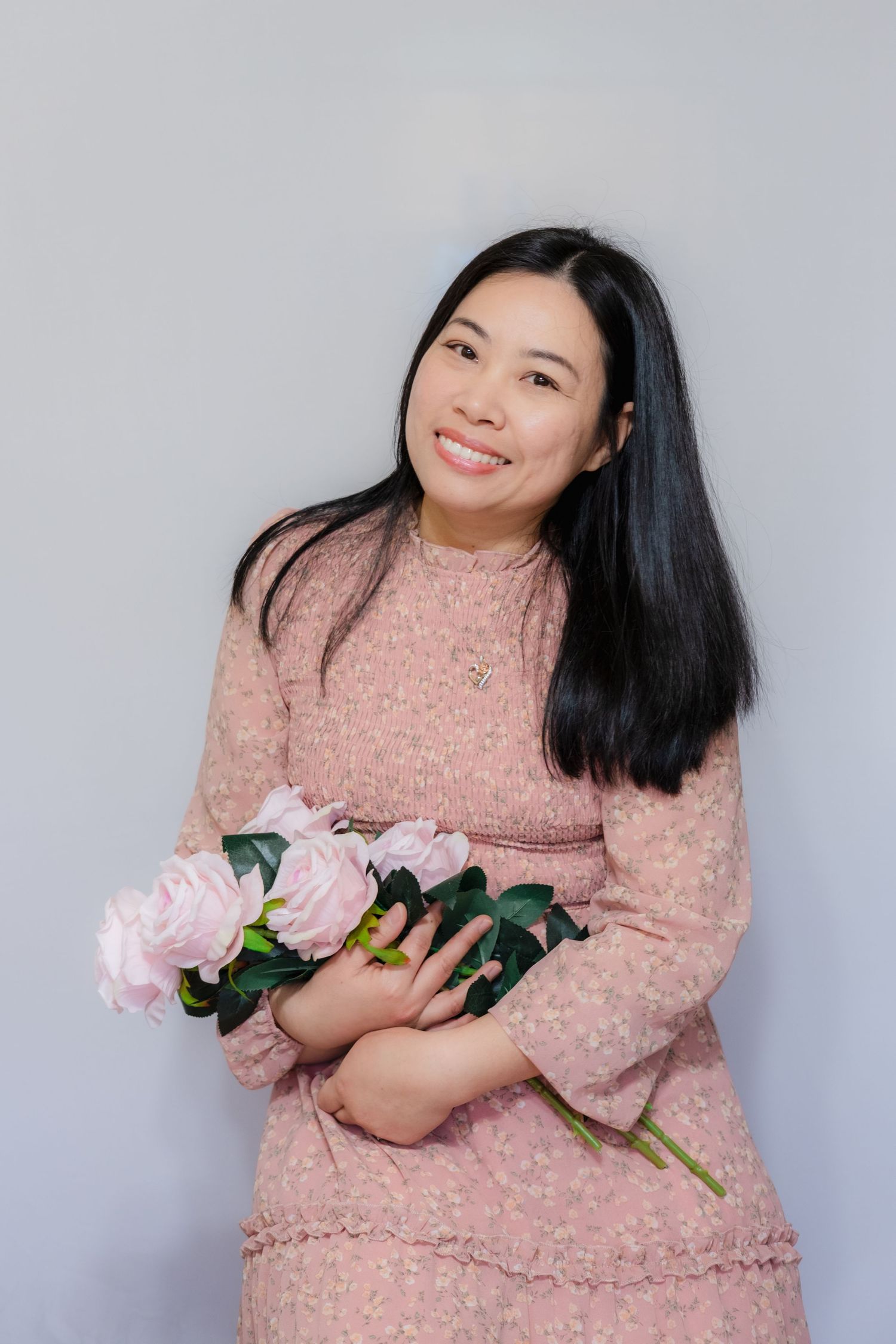 Smiling Asian woman in pink floral dress holding light pink peonies against a soft gray background.
