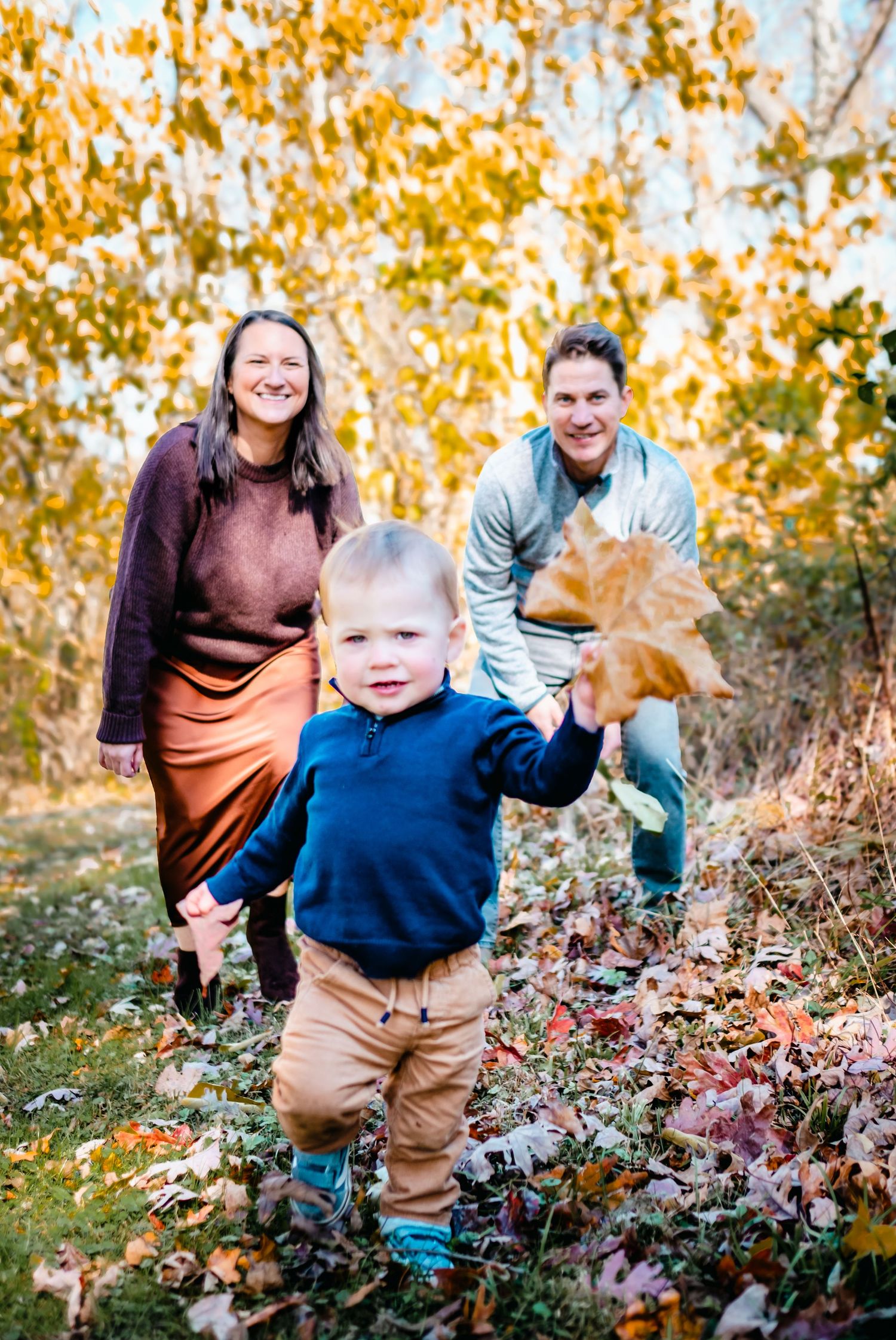 Family of three enjoying autumn outdoors, toddler holding a leaf while parents smile among colorful fall foliage.