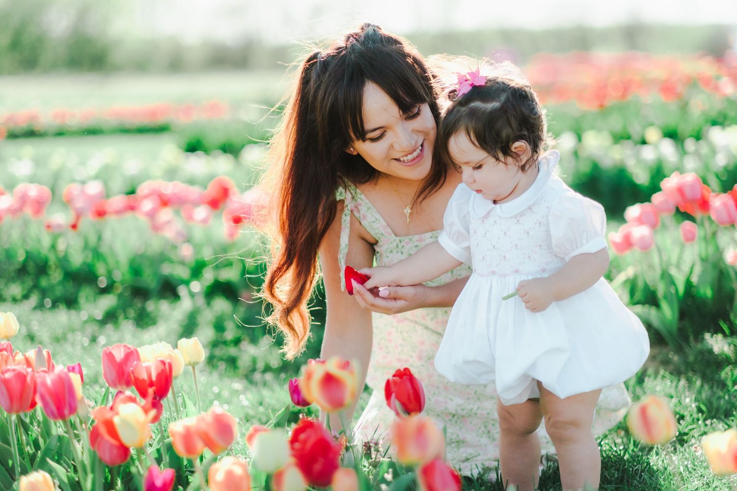 A smiling mother and baby daughter in white dress admiring colorful tulips in a vibrant spring flower field.