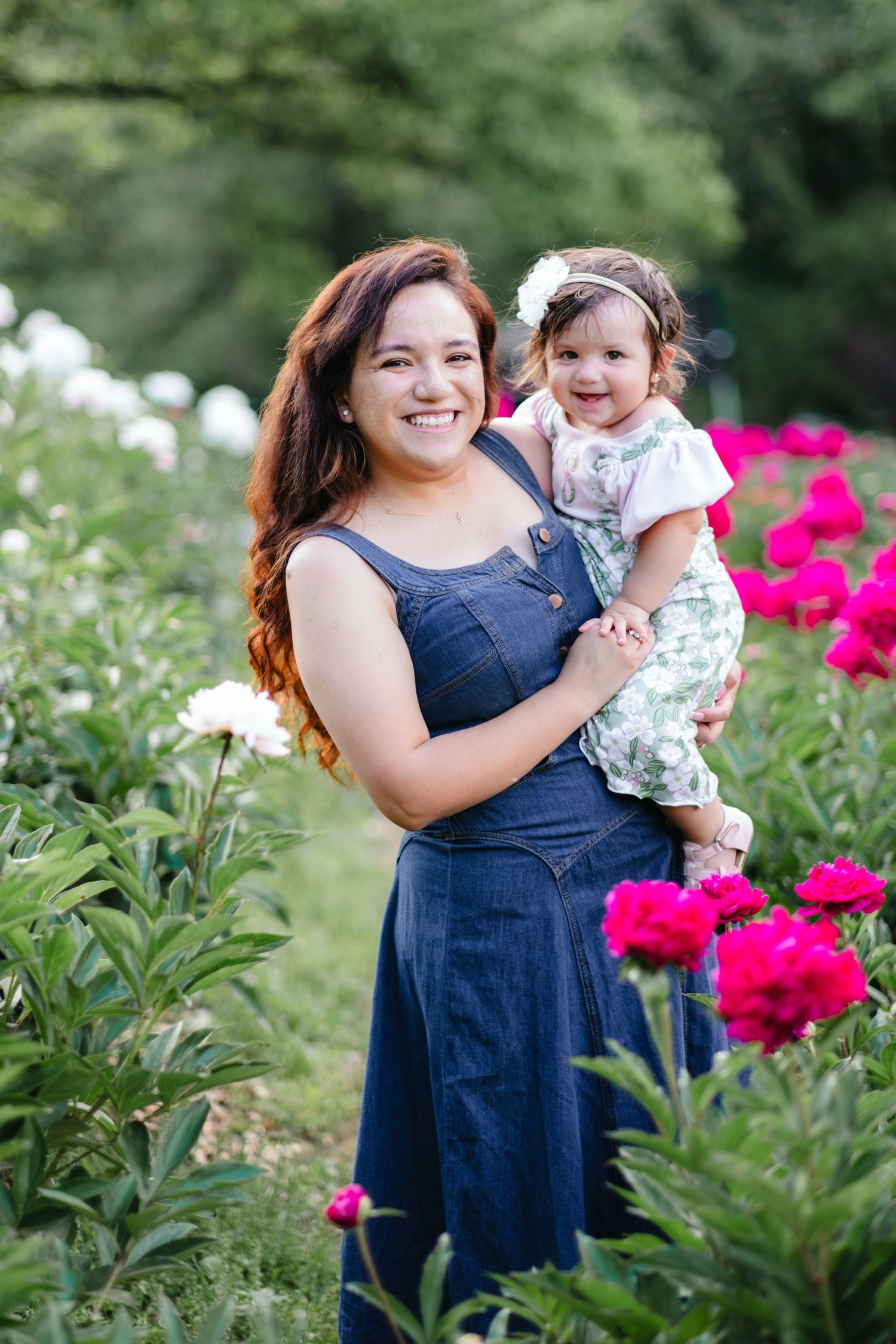 A smiling woman in a denim dress holds a baby girl among vibrant pink and white peony flowers in a lush garden.