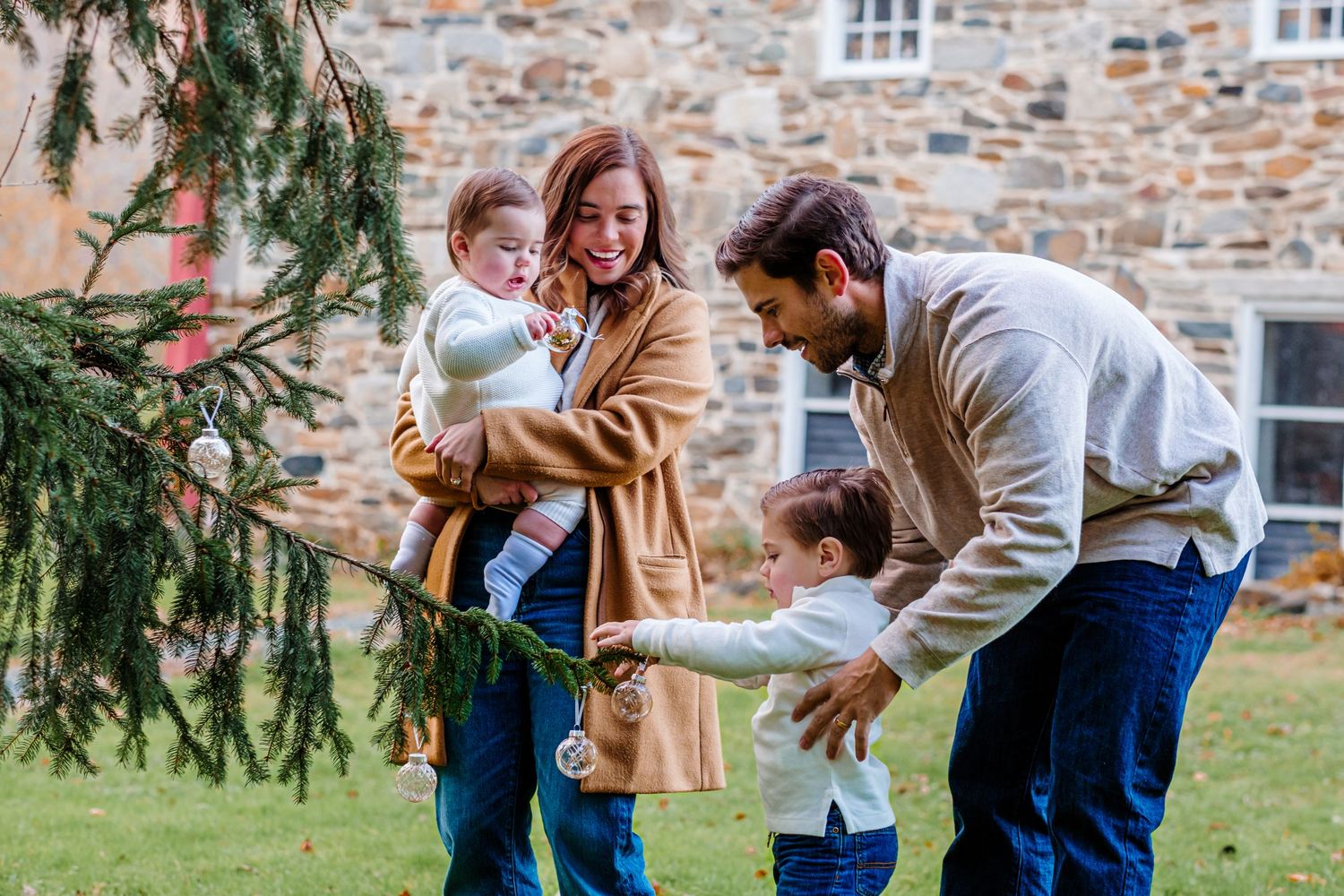 Family of four decorating an outdoor Christmas tree together on a green lawn beside a stone building.