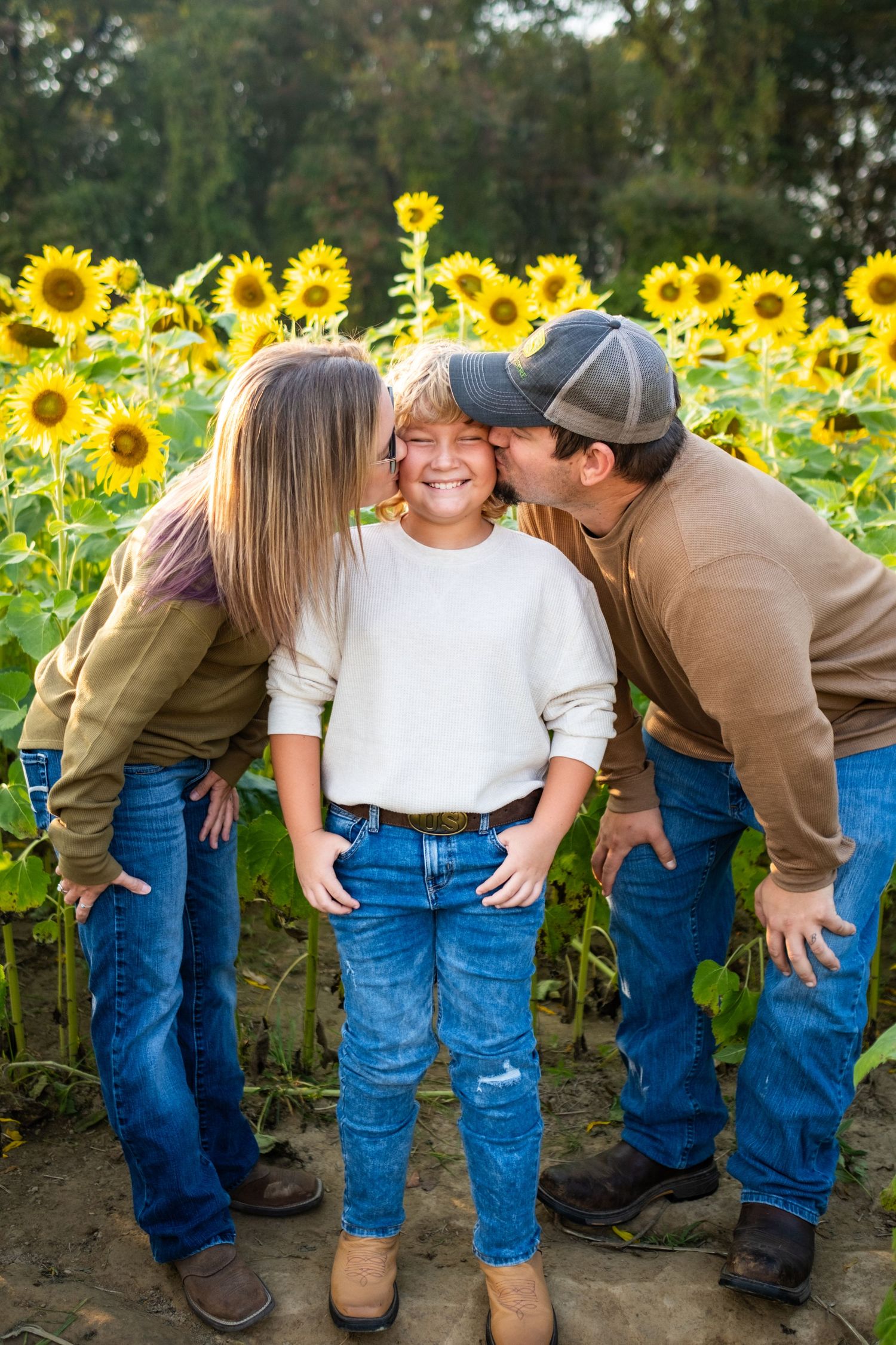 Parents kissing their smiling child on the cheeks in a vibrant sunflower field, family wearing jeans and casual tops.