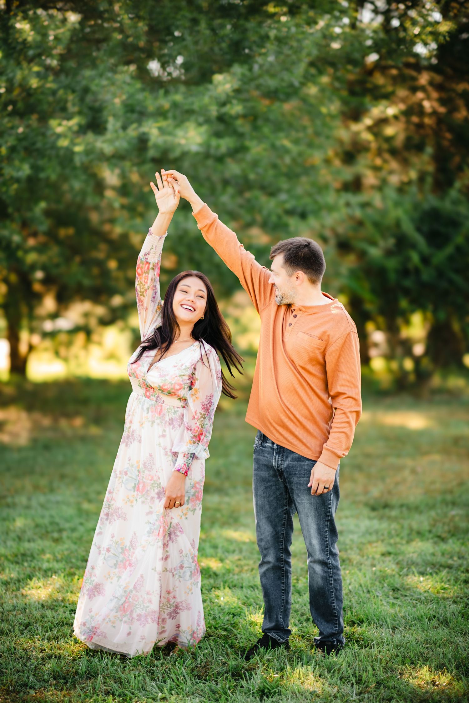 A joyful couple dancing outdoors, woman in floral dress being twirled by man in orange shirt amid lush green trees.