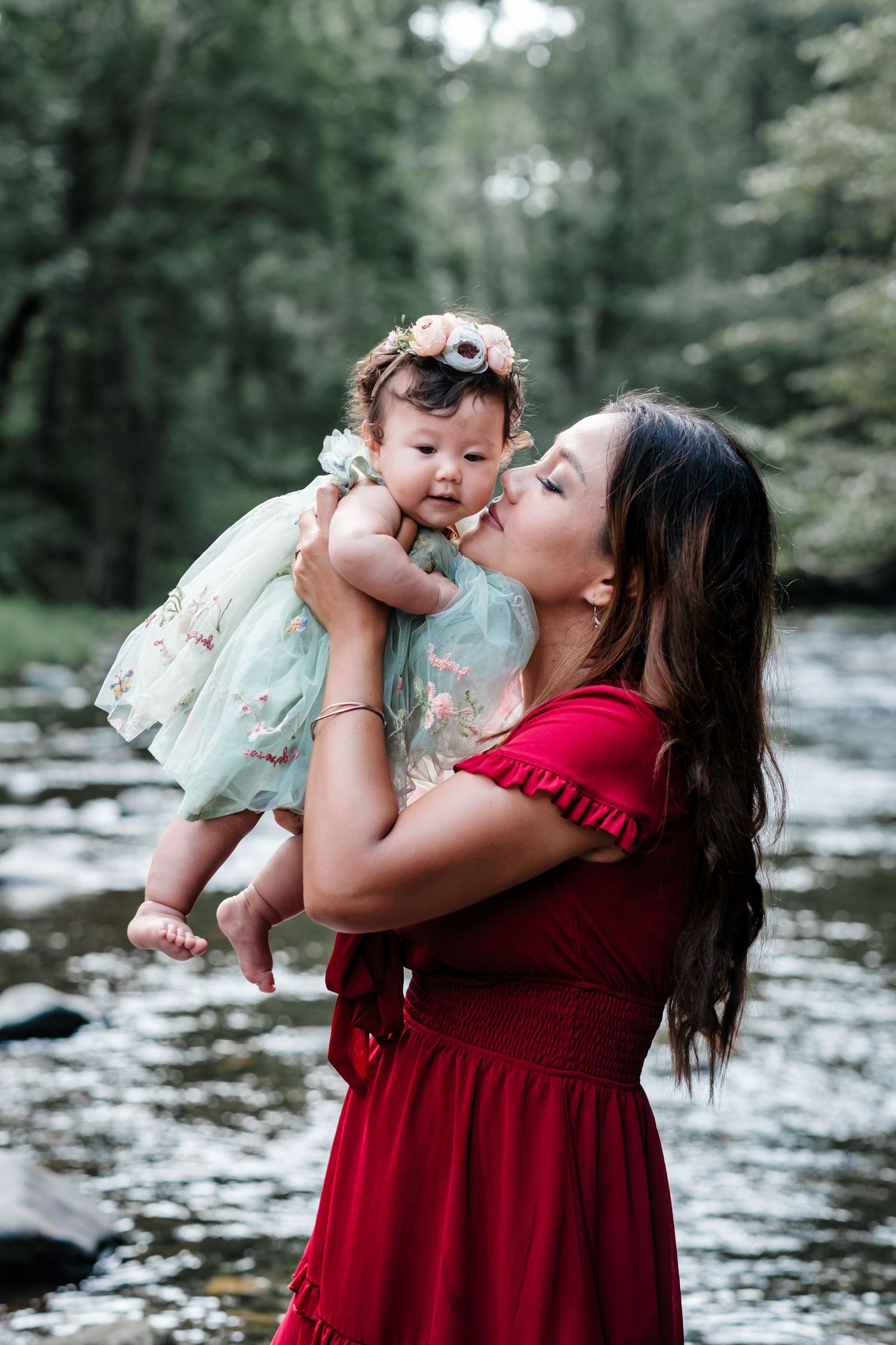 A mother in a red dress kisses her baby girl in a floral dress by a serene forest creek.