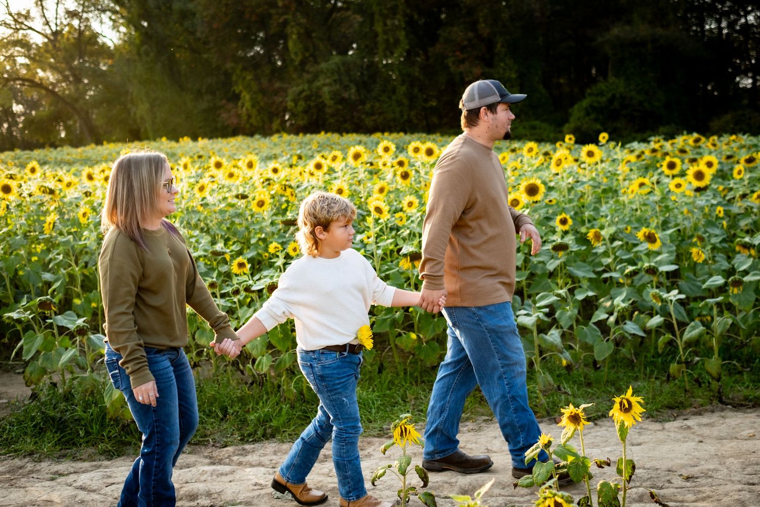 Family of three walking hand-in-hand through a vibrant sunflower field at golden hour.