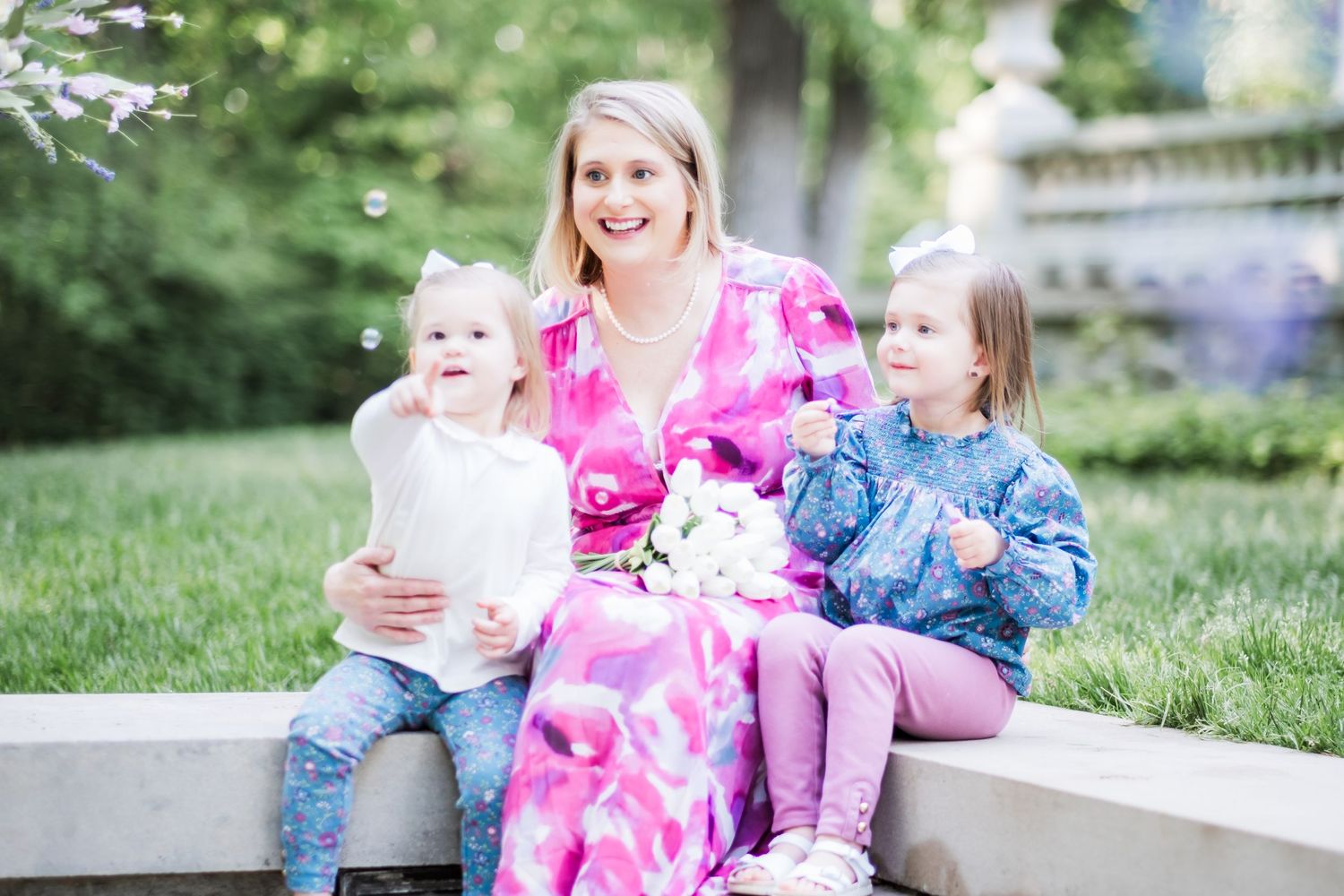 Three people sit together on a stone ledge in a garden setting wearing casual clothing and pink floral patterns.