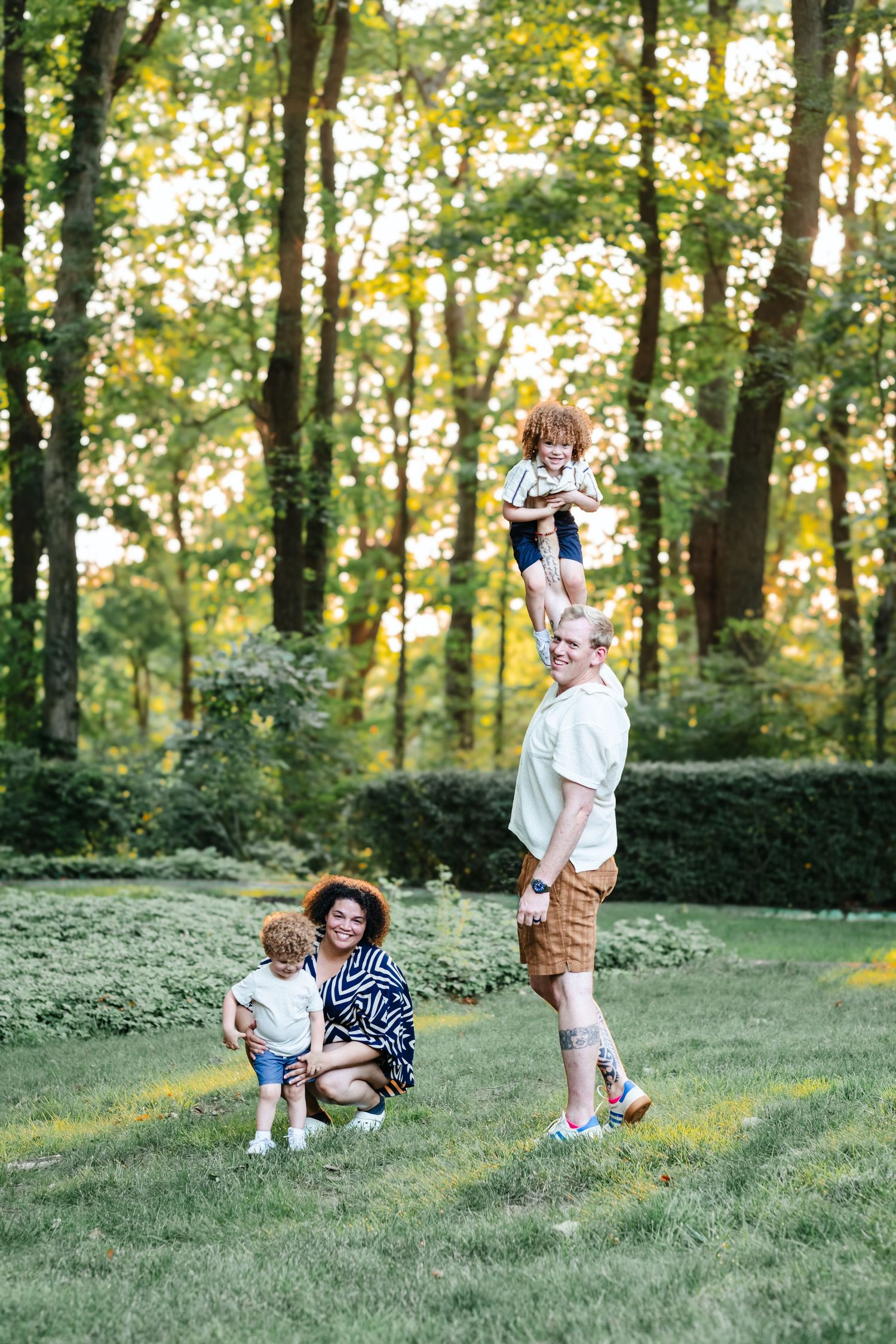 Family playing outdoors in lush green park, father lifting child overhead while mother crouches with toddler on sunny day.