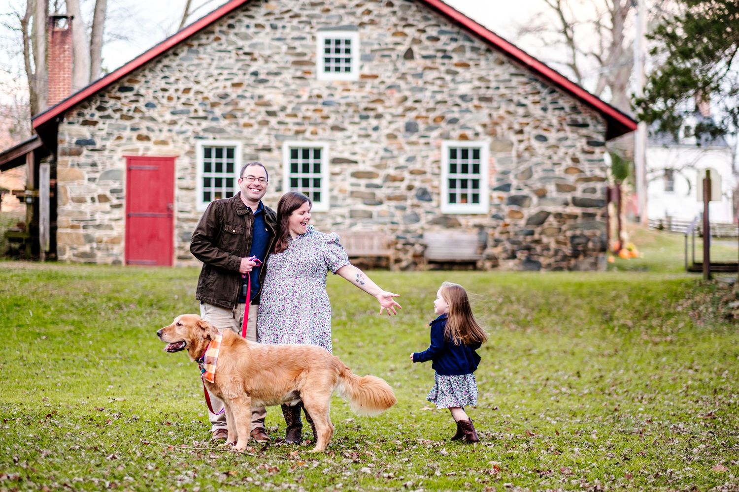 Family with two golden retrievers laughing outside a rustic stone barn with a red door on a green lawn.