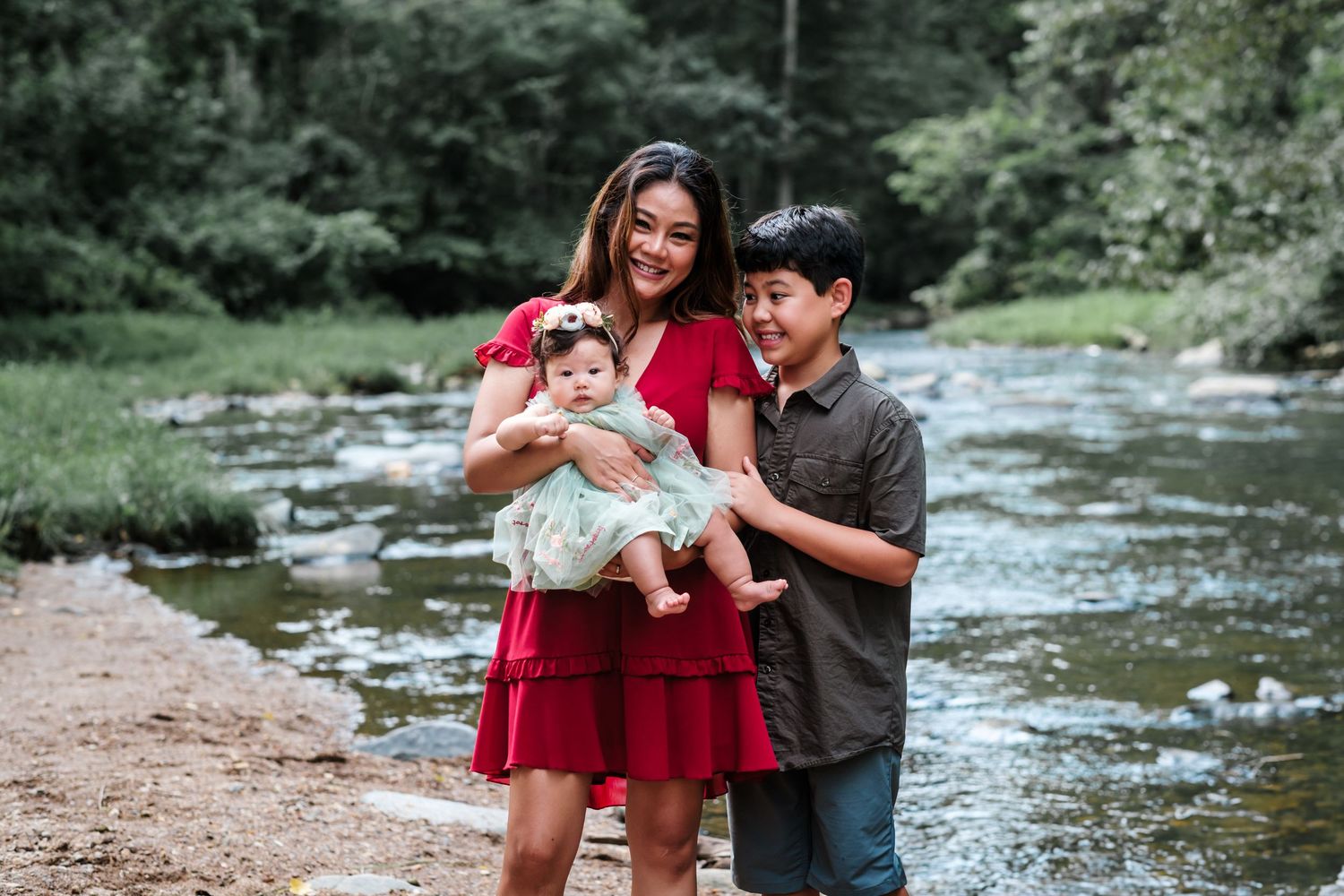 A woman in a red dress holds a baby girl while a young boy stands beside them near a scenic creek.
