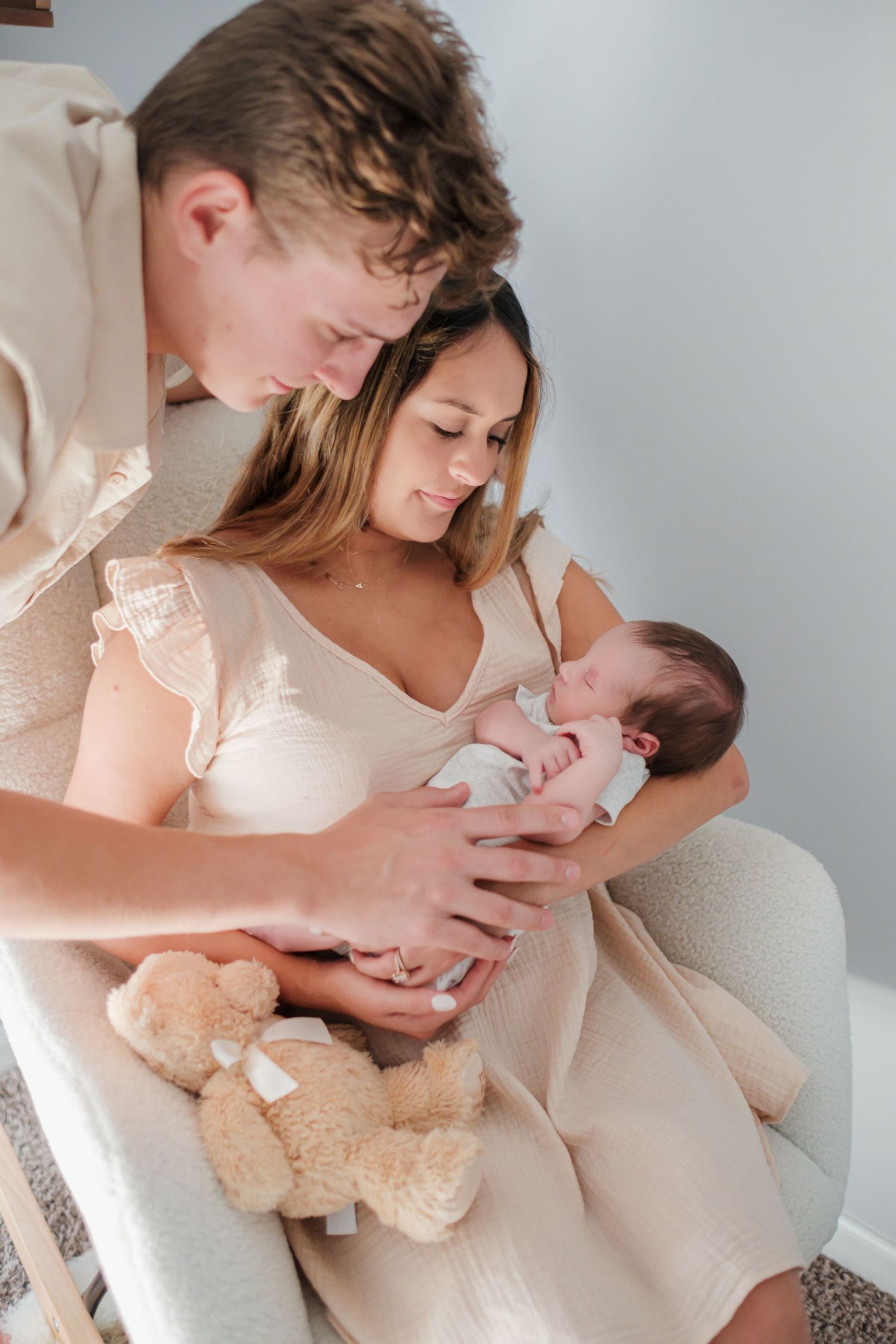 Young couple in beige outfits lovingly gazing at their newborn baby while holding a teddy bear indoors.