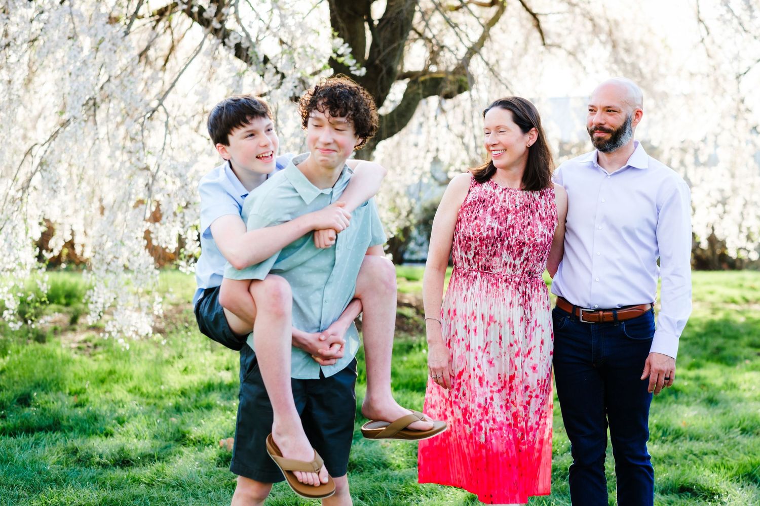 Family of four smiling outdoors near blooming white cherry blossom tree, two boys and parents on green lawn.