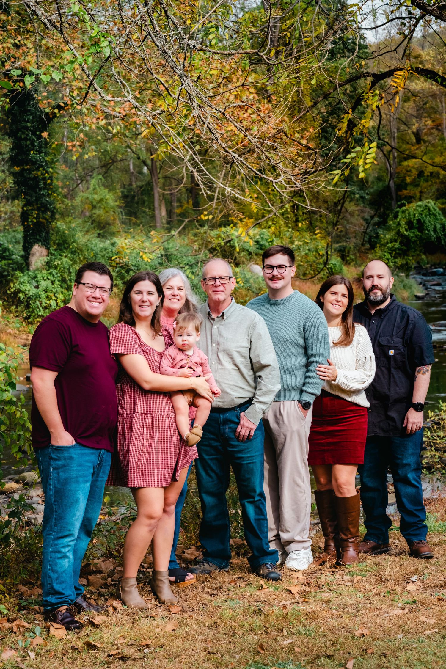 Family of seven poses outdoors in autumn, surrounded by colorful fall foliage near a creek.