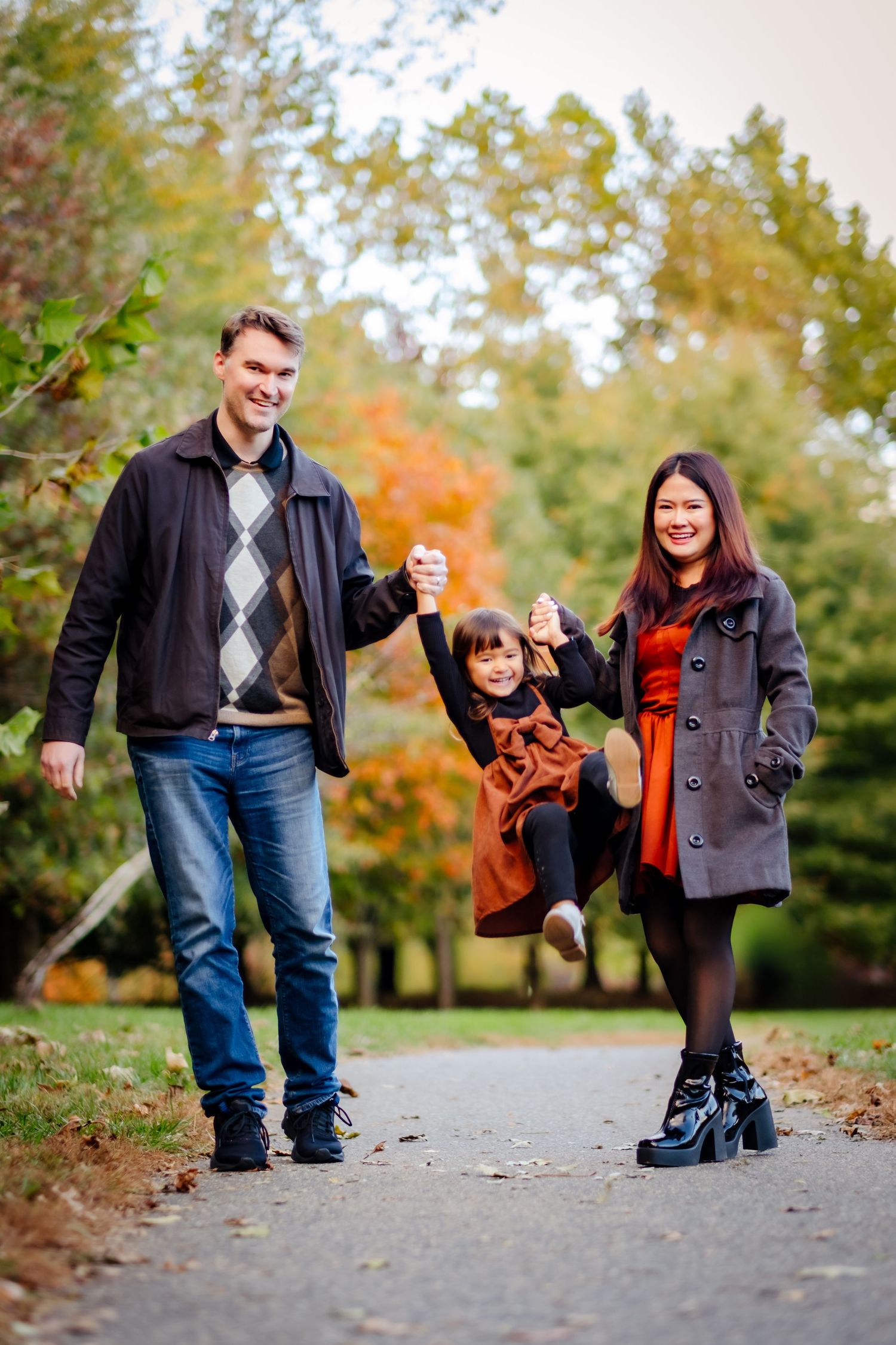 Family of three enjoying autumn walk in park, parents swinging young child between them on tree-lined path.