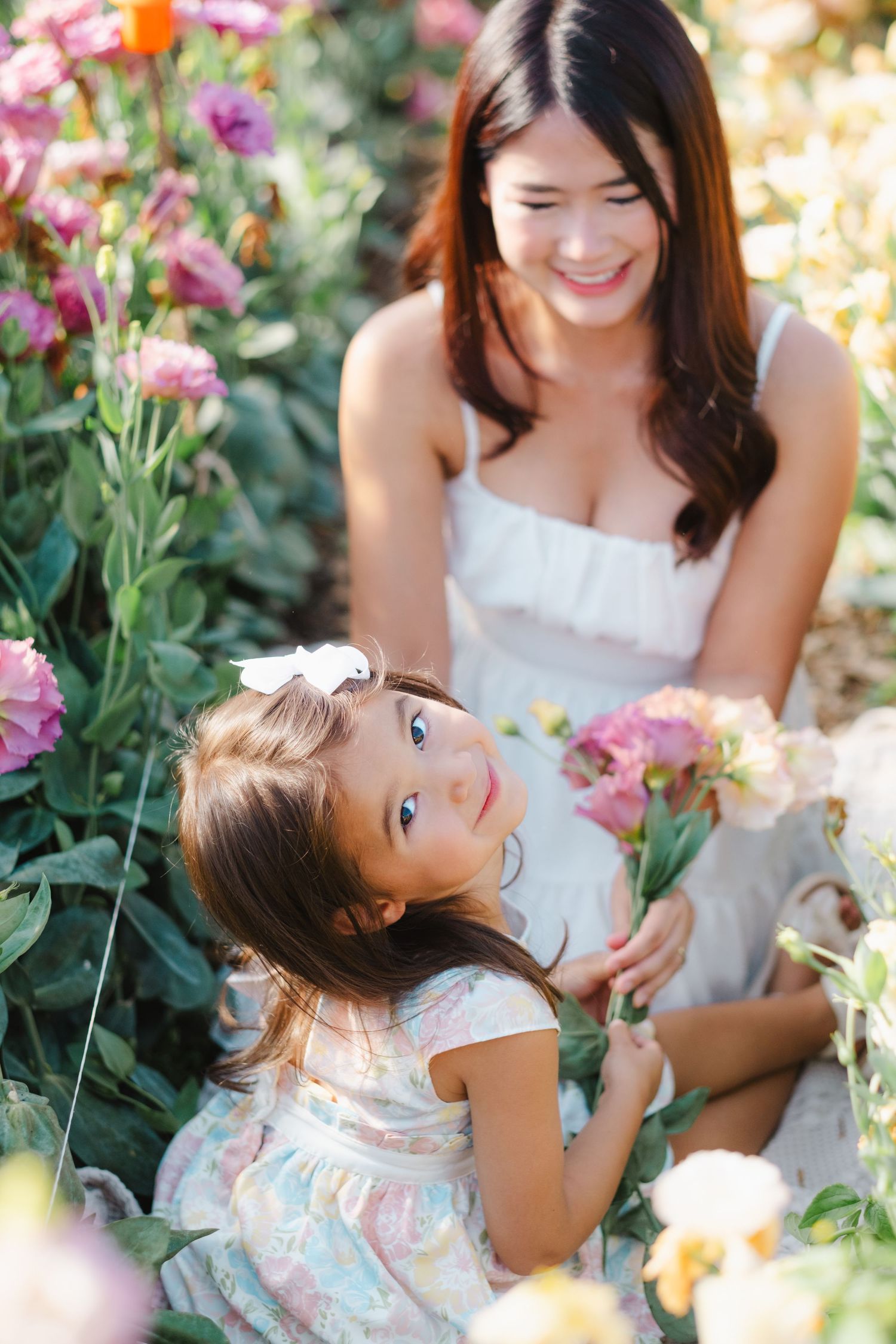 A smiling woman and young girl in white dresses sit among colorful blooming flowers in a sunny garden.