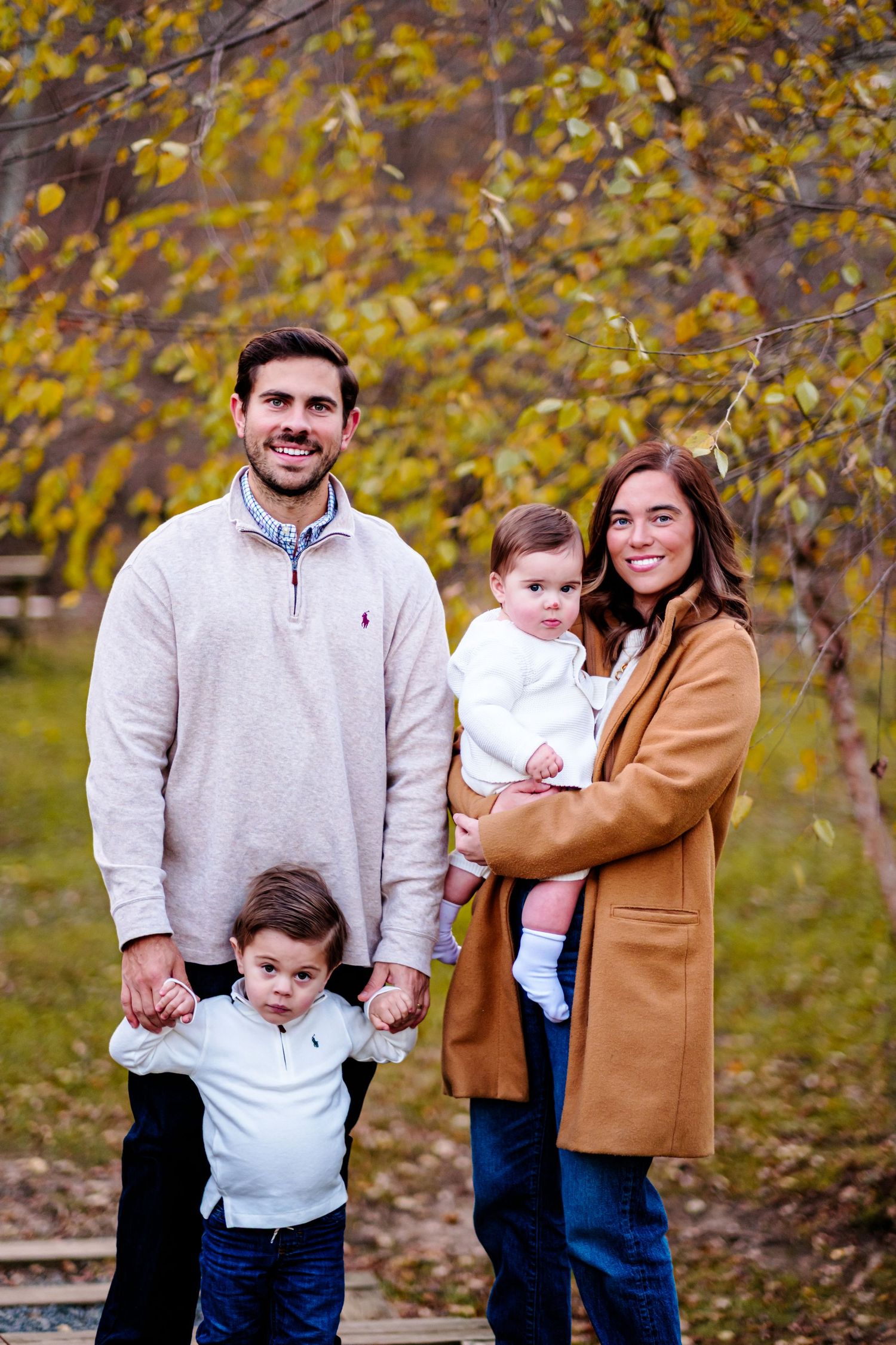 A smiling family of four poses outdoors in autumn, parents holding two young children among golden fall foliage.
