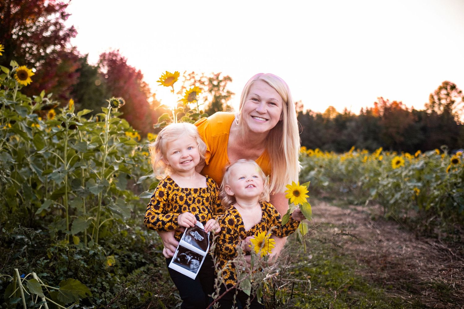 Woman in yellow top poses with two young girls in sunflower dresses in a sunlit sunflower field.
