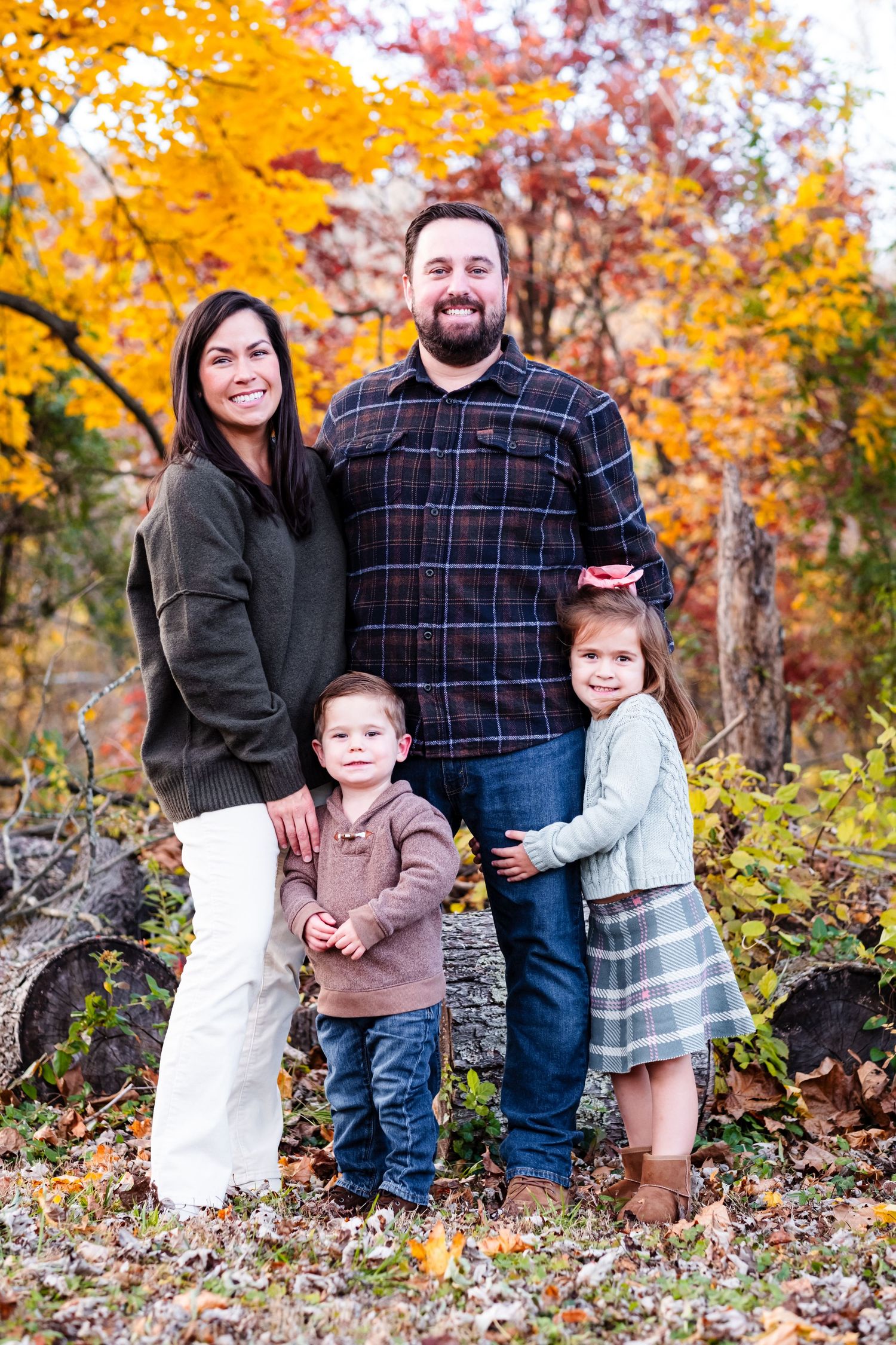 Family of four posing outdoors among vibrant fall foliage, parents with two young children smiling in autumn setting.