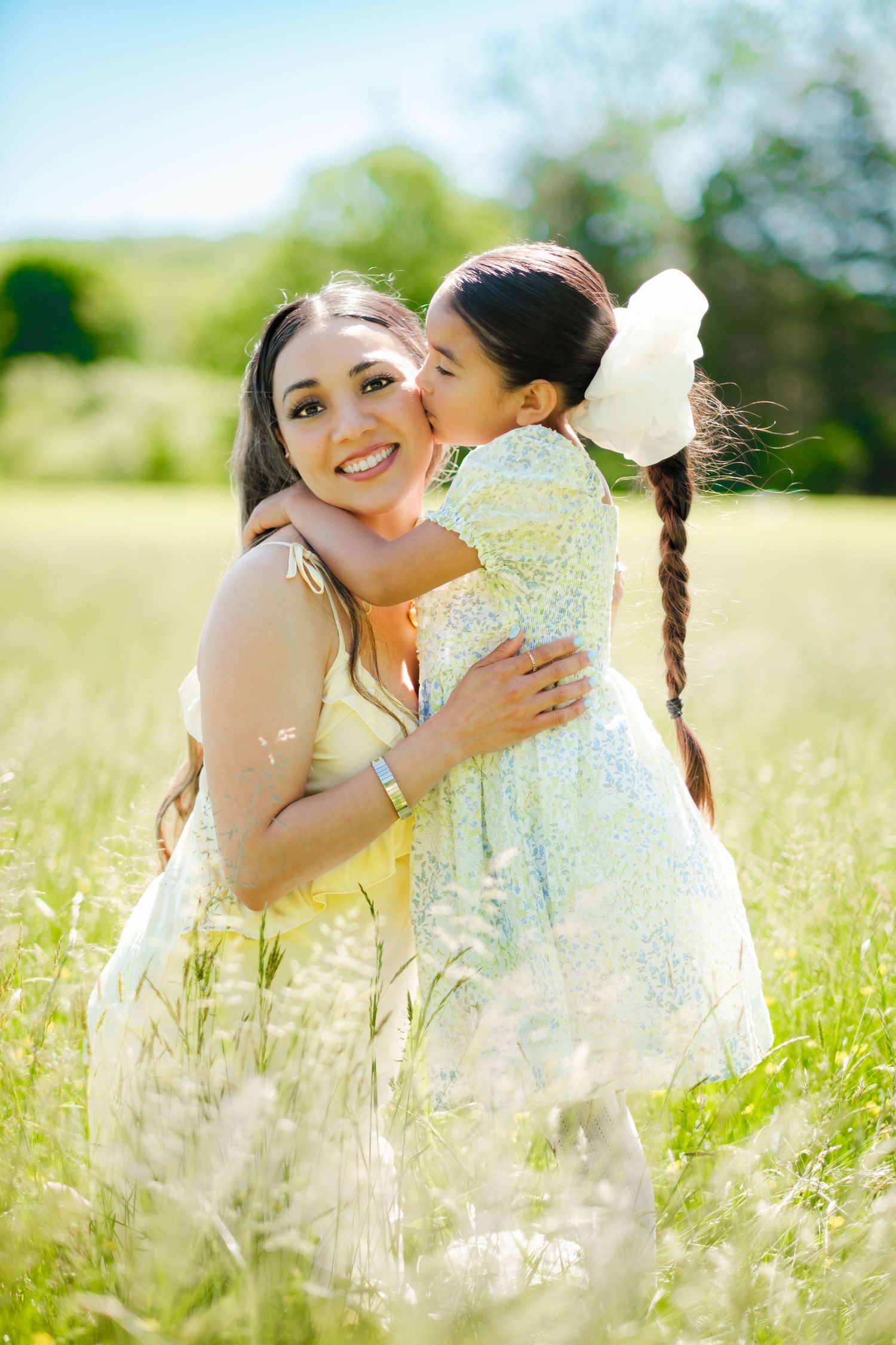 A smiling woman in a yellow dress hugs a girl in a floral dress sharing a kiss in a sunny green field.