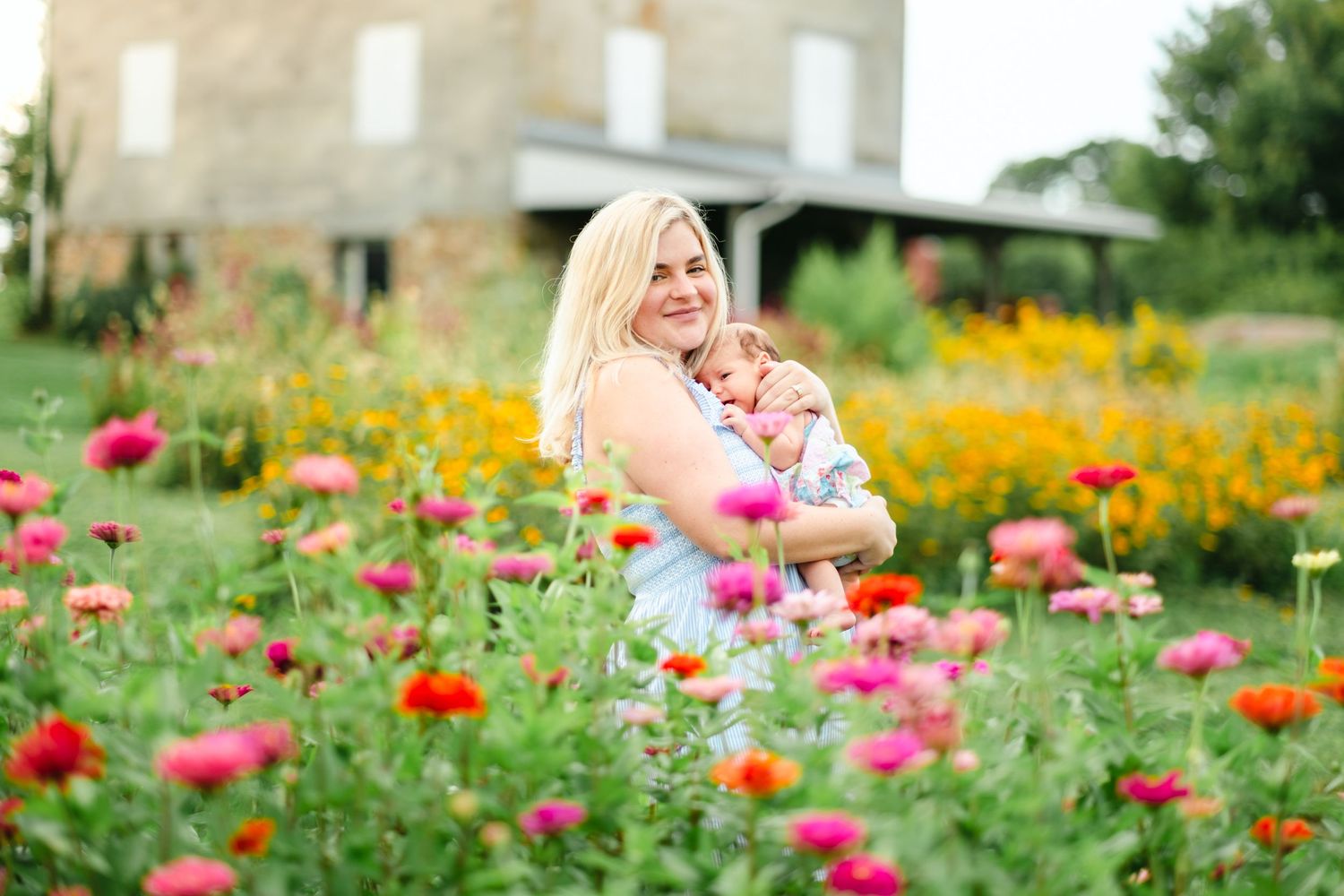 Blonde woman holding a baby in a vibrant flower field with pink, red, and yellow blooms near a stone building.