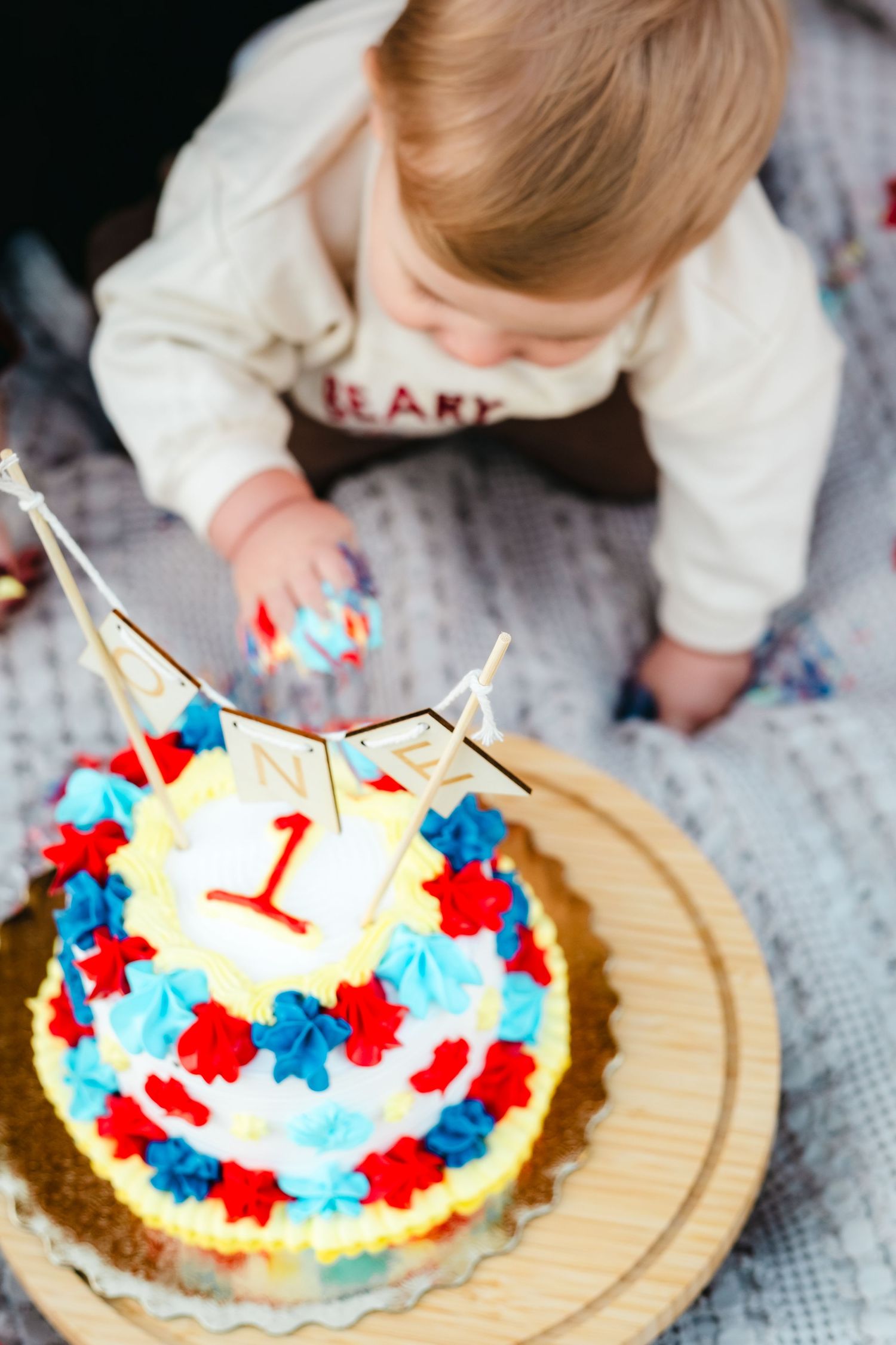 Baby reaching toward a colorful first birthday cake decorated with red and blue flowers and a number 1 topper.