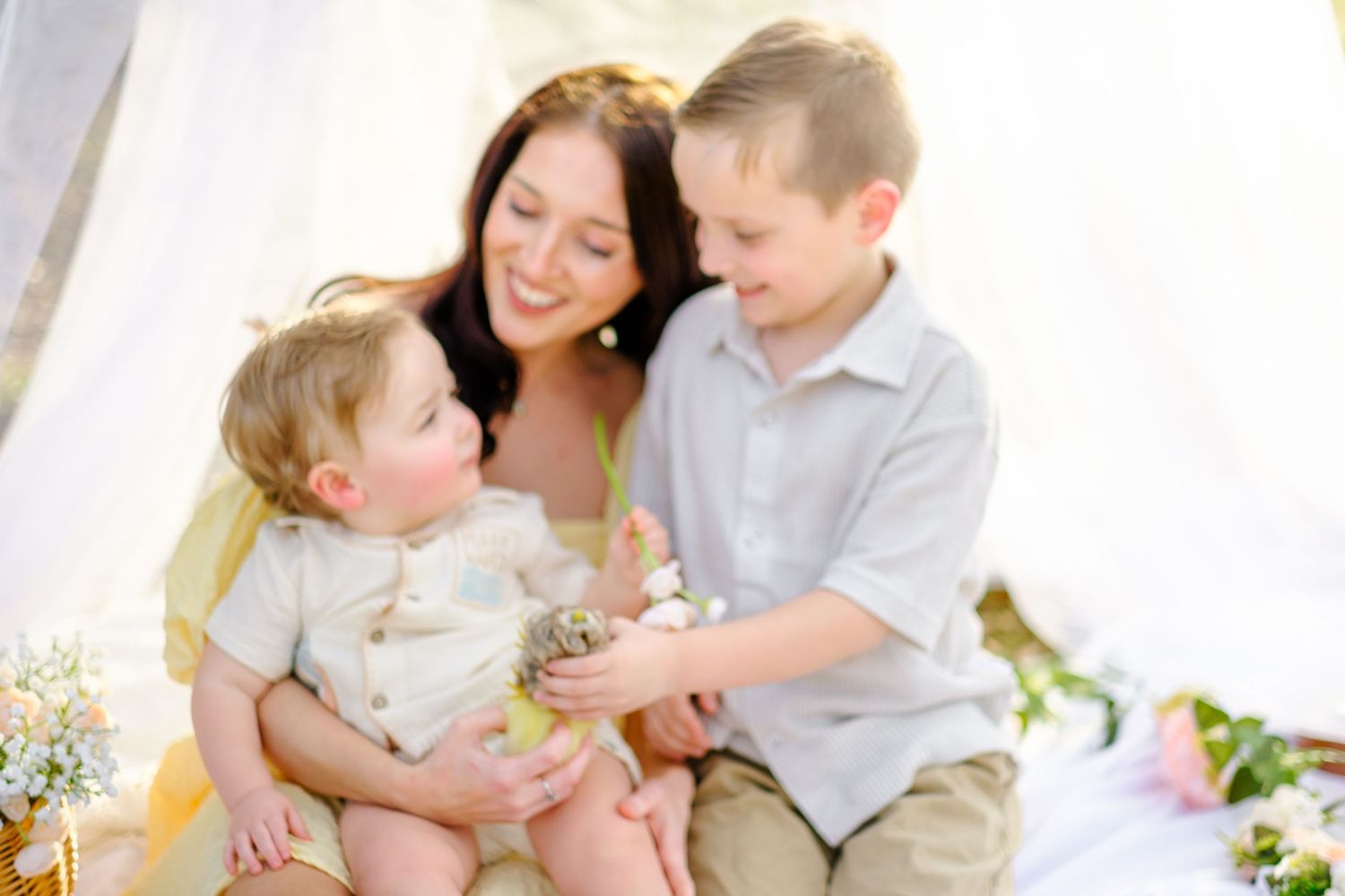 A mother smiles warmly while holding her toddler as an older boy shows them a small nest outdoors.