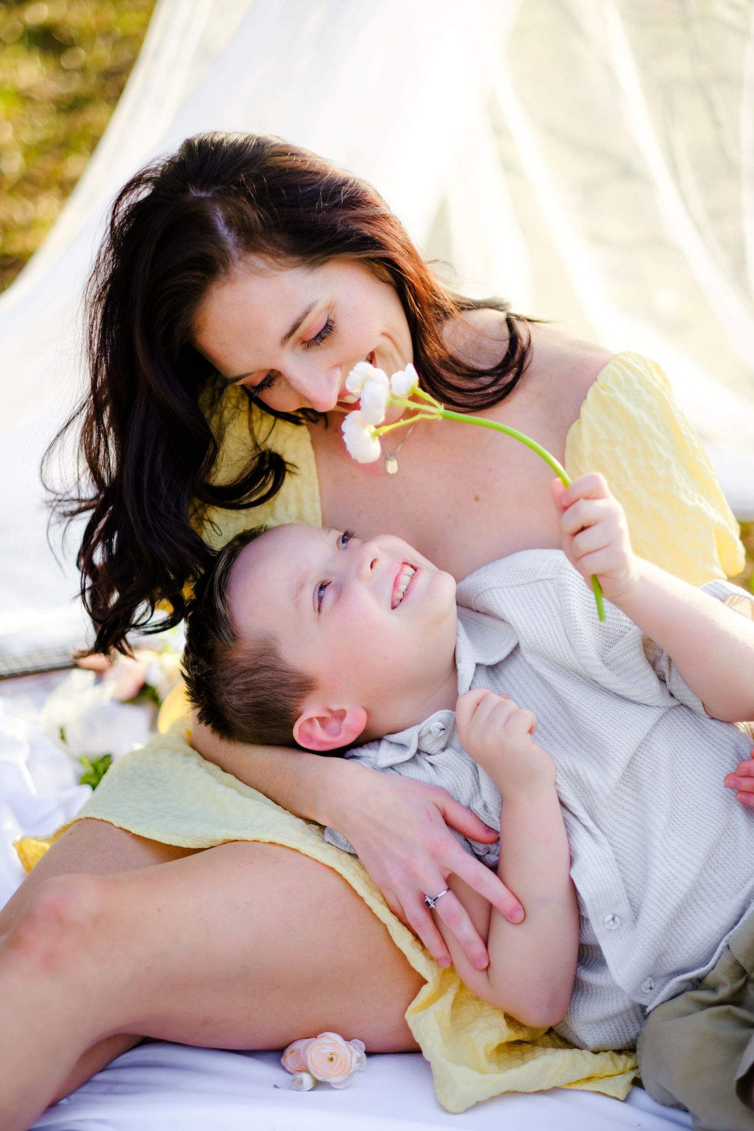 A mother and young son share a tender moment outdoors, as she holds a white flower while he smiles up at her.
