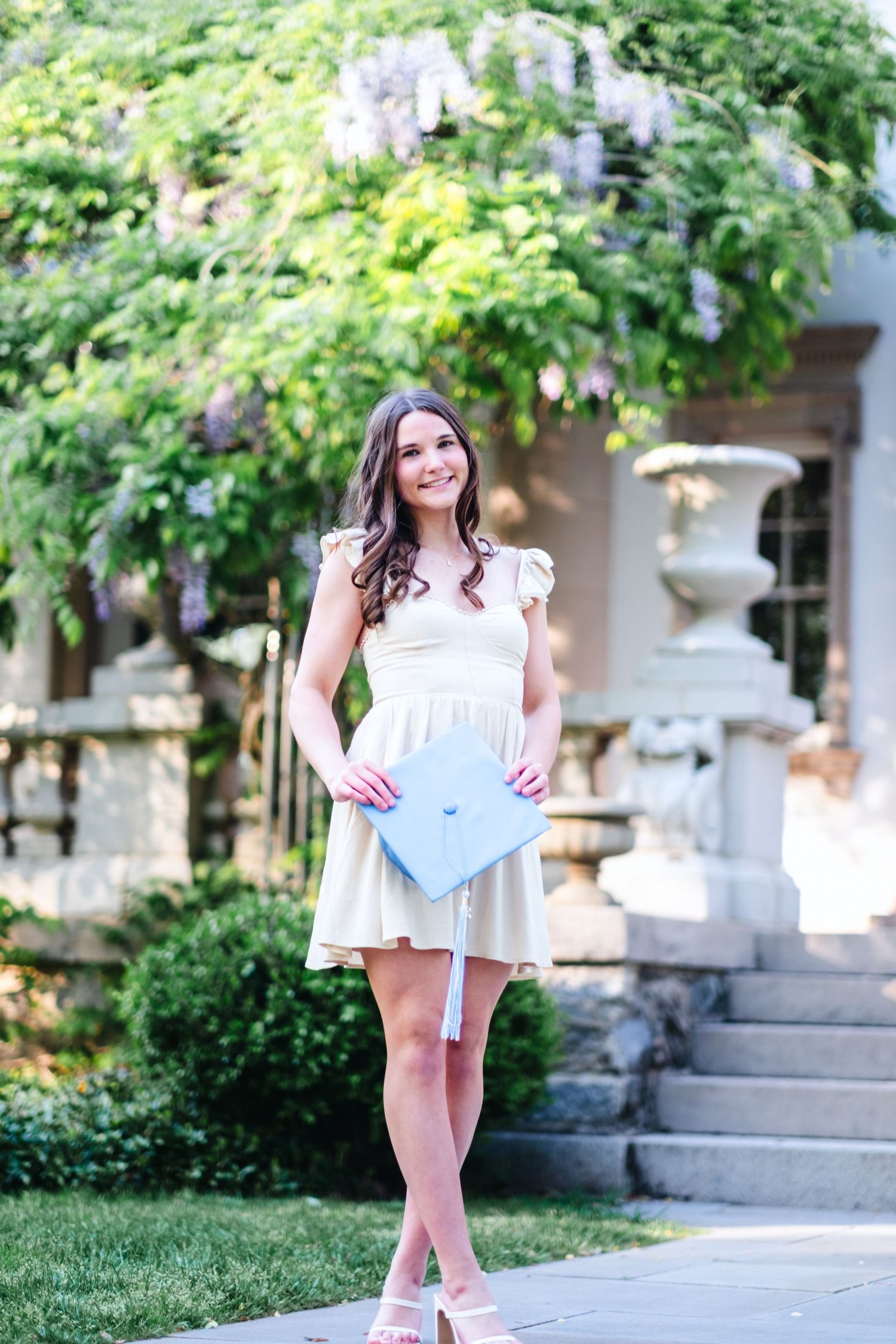 Young woman in cream dress holding graduation cap outdoors near stone fountain and blooming wisteria.